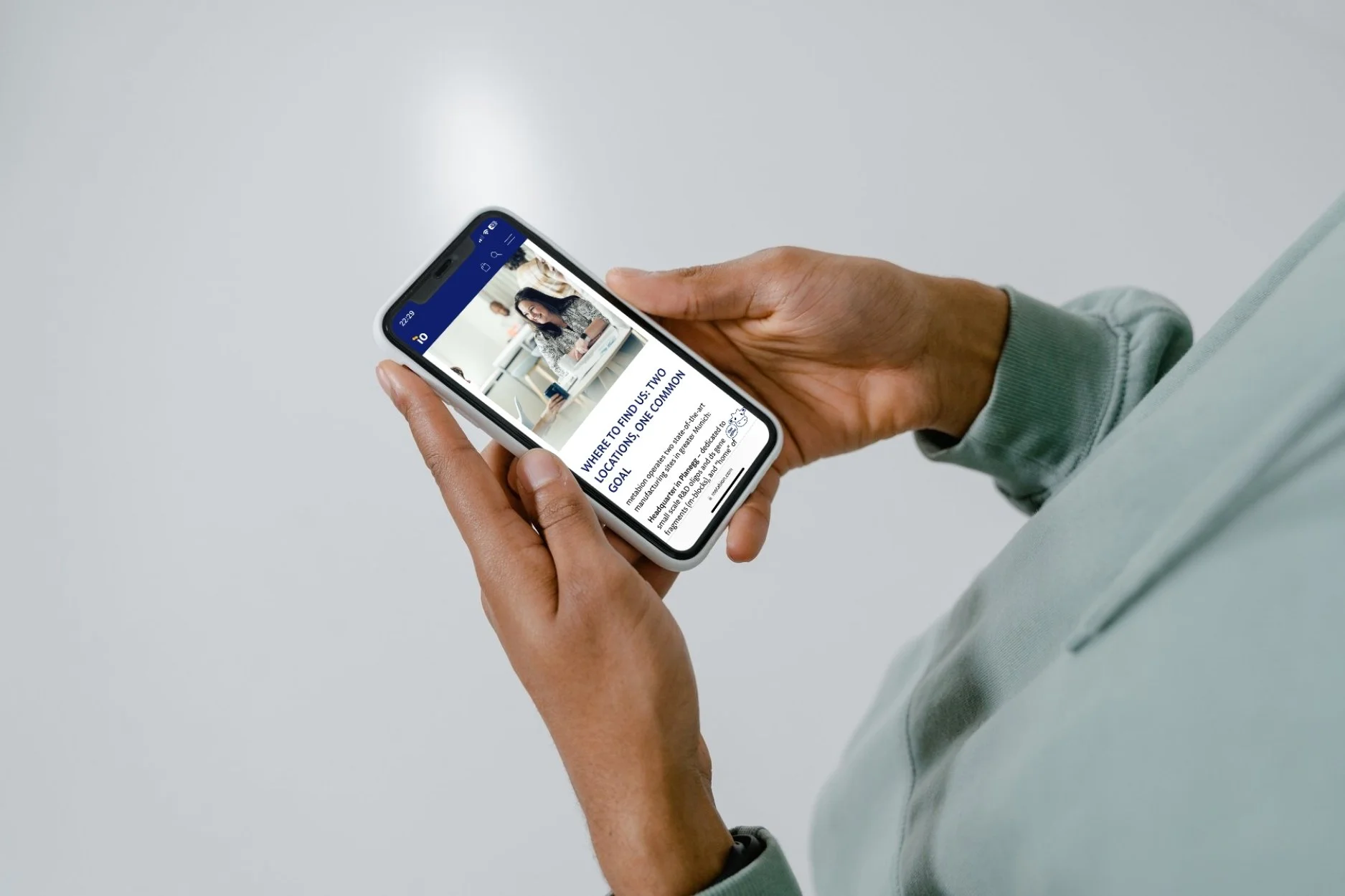 Nahaufnahme einer Person, die ein Smartphone hält und eine Webseite auf dem Bildschirm liest. Die Person trägt eine graue Sweatjacke und hat dunkle Haut.