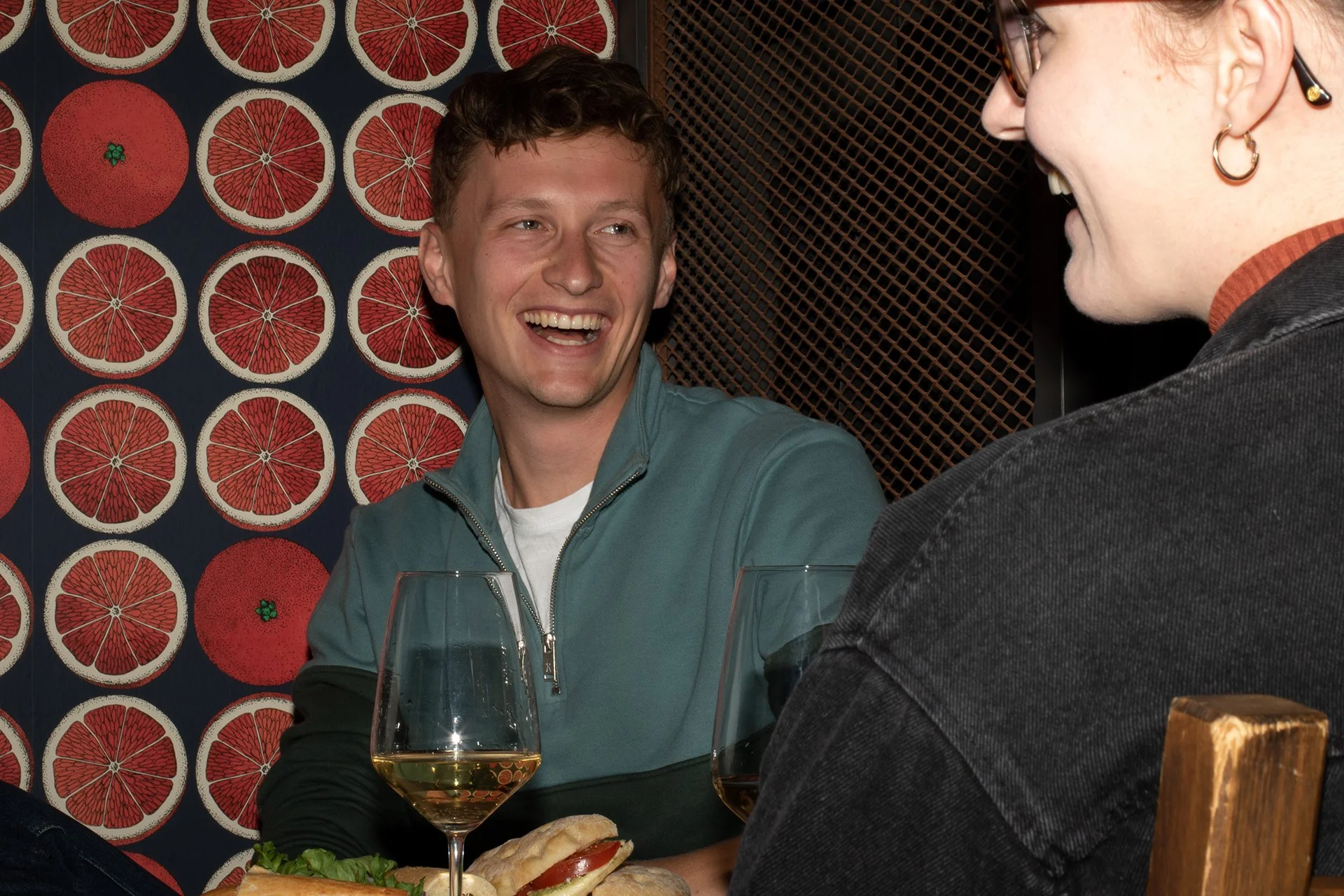 Zwei Menschen sitzen an einem Tisch, lachen und unterhalten sich. Im Vordergrund stehen zwei Gläser Weißwein, und auf dem Tisch liegt ein Sandwich mit Salat, Tomate und Frischbrot. Die Wand im Hintergrund ist mit einem Muster aus Grapefruit-Scheiben dekoriert.