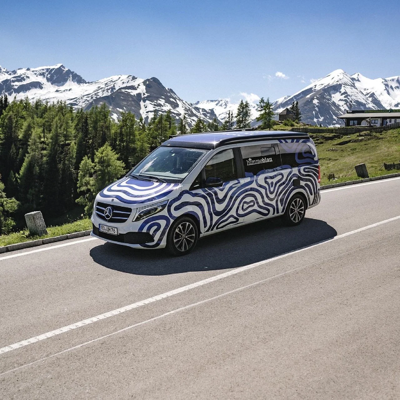 Ein schwarzer Mercedes-Benz Transporter mit blauen geometrischen Mustern, der auf einer Bergstraße im Alpengebiet steht, mit Schneebedeckten Bergen im Hintergrund und grünen Wäldern.