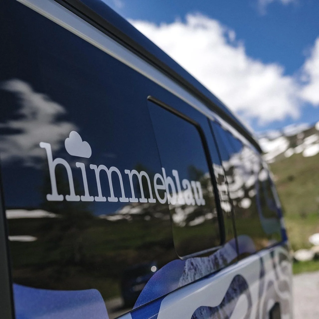 Aufkleber mit dem Schriftzug 'himmeblau' und einer Wolke auf einer dunklen Fahrzeugseite, mit Berg und Himmel im Hintergrund.