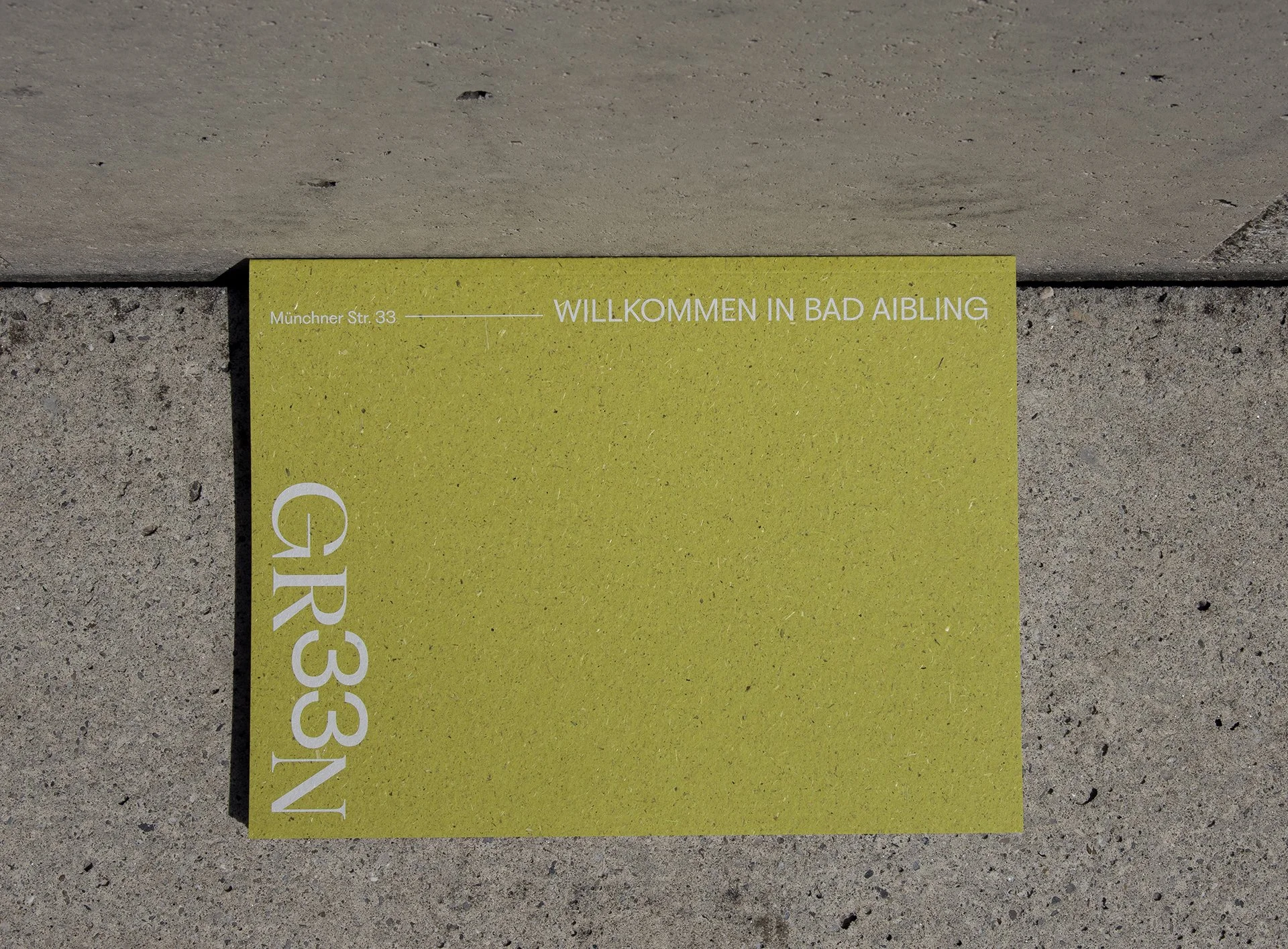 Ein grünes Schild mit weißen Schriftzug, auf dem steht: 'Münchner Str. 33, WILLKOMMEN IN BAD AIBLING', auf einem Betonboden liegend.