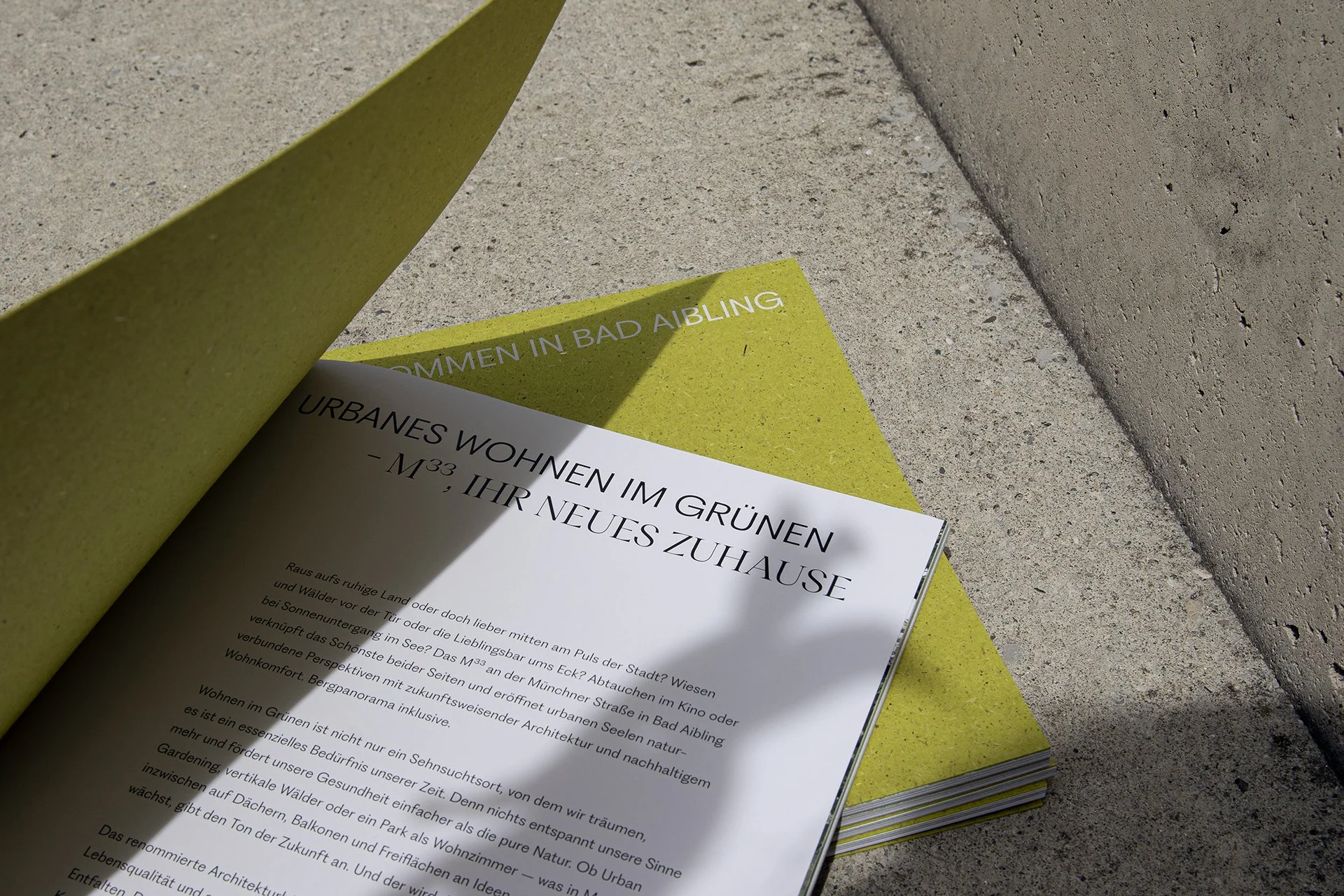 Offene Broschüre mit Text auf deutschen Seiten, liegt auf einer gelben Unterlage auf einer Betonoberfläche. Der sichtbare Text enthält den Titel „URBANES WOHNEN IM GRÜNEN – M33, IHR NEUES ZUHAUSE“.