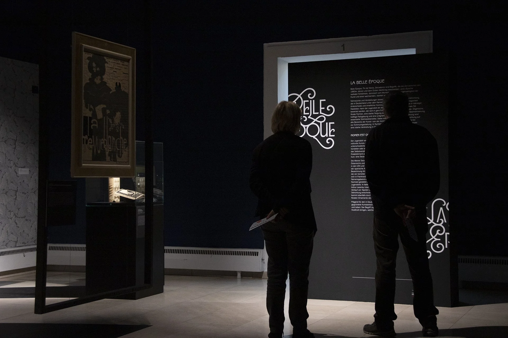Zwei Menschen schauen auf ein hell beleuchtetes Informationstablett in einem dunklen Museum. Weitere Ausstellungsstücke sind zu sehen, darunter ein gerahmtes Plakat an der Wand.