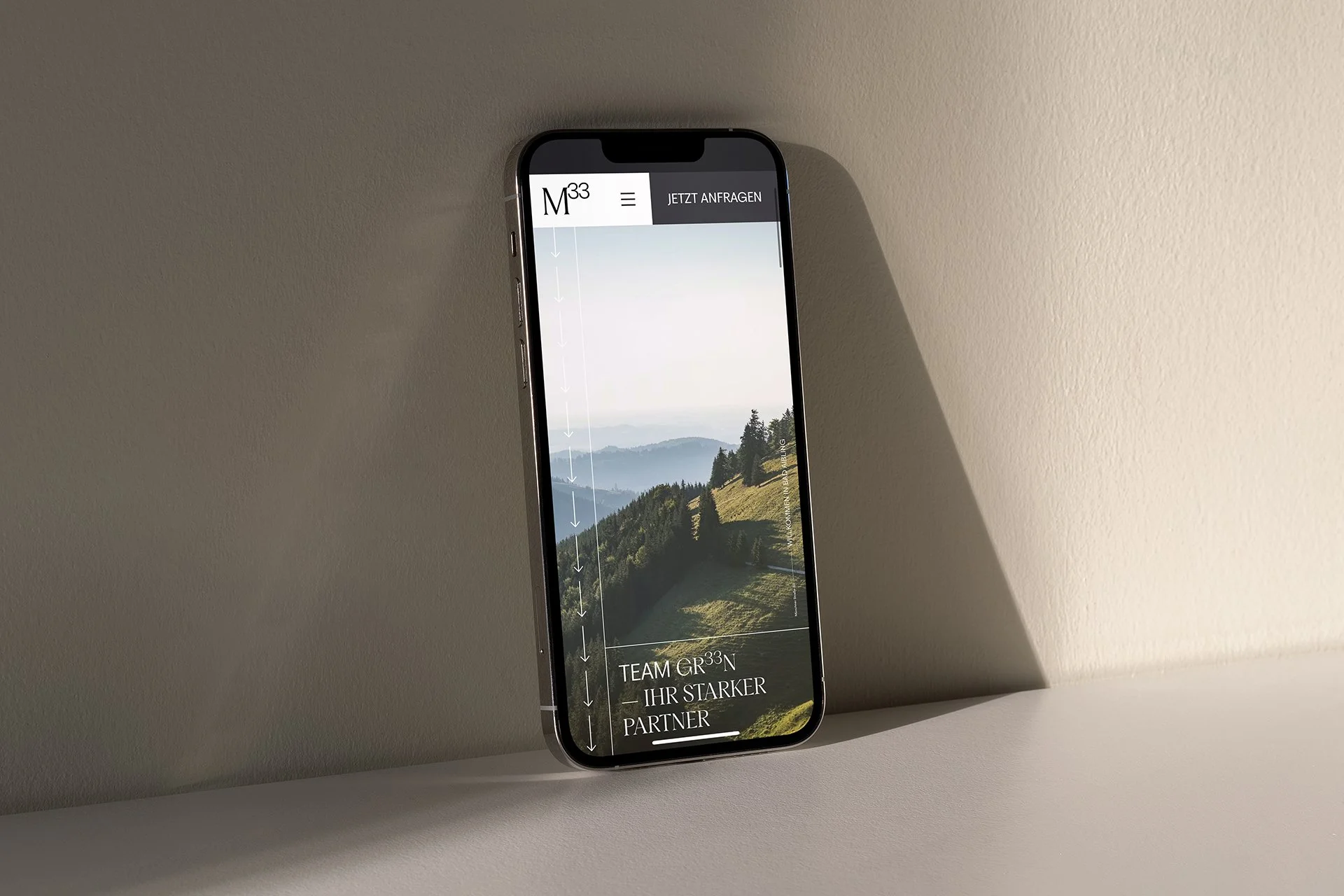 Smartphone auf einem weißen Tisch, zeigt die Webseite eines Unternehmens namens M33 mit einem Wandbild eines Berg- und Wiesenhorizonts und Text, der auf Deutsch für Teamarbeit und starke Partnerschaften wirbt.