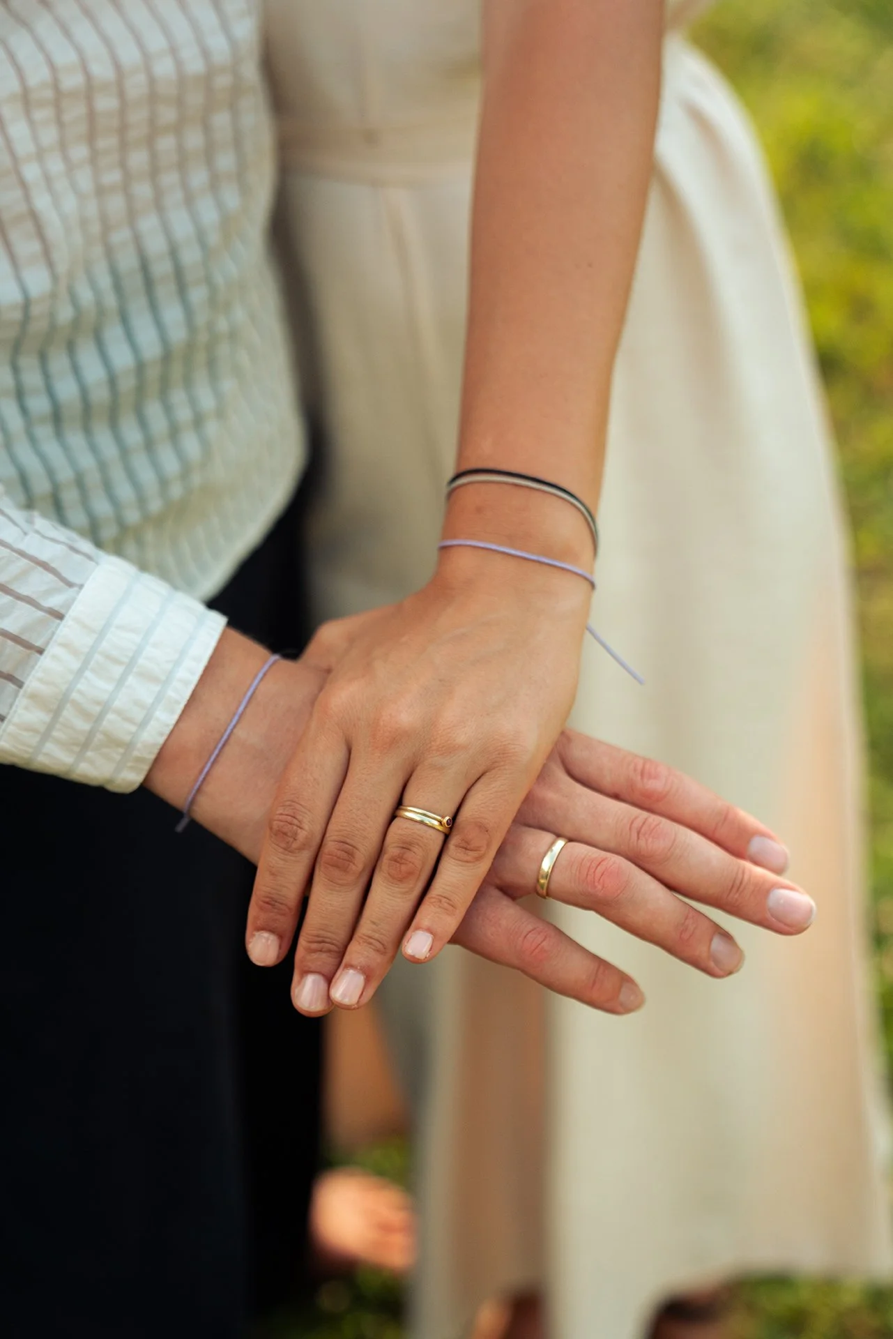 Zwei Personen berühren sich mit verschränkten Händen und tragen Ringe. Eine Person trägt mehrere Armbänder und eine längere, gestreifte Bluse. Die andere Person trägt eine schwarze Hose.
