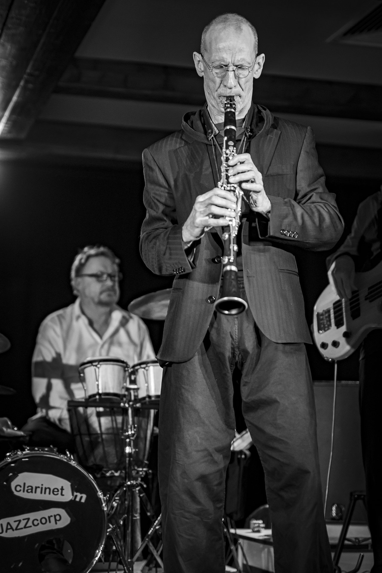 Ein älterer Mann spielt auf einem schwarzen Klarinette, im Hintergrund sitzt ein Schlagzeuger und eine Gitarre ist sichtbar. Es ist eine Jazz-Performance, Szene in Schwarz-Weiß.