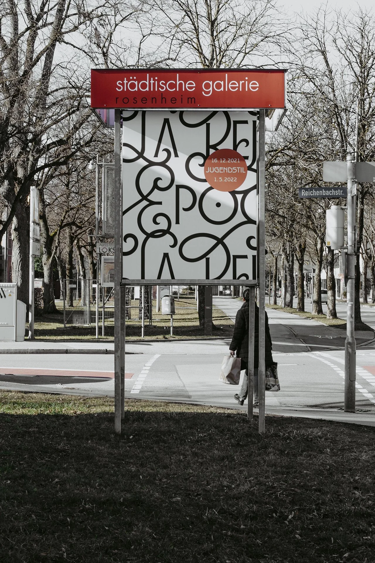 Straßenschild mit der Bezeichnung Reichenbachstraße, ein Plakat mit Kunst im öffentlichen Raum, und eine Person, die eine Einkaufstasche trägt, während sie die Straße überquert.
