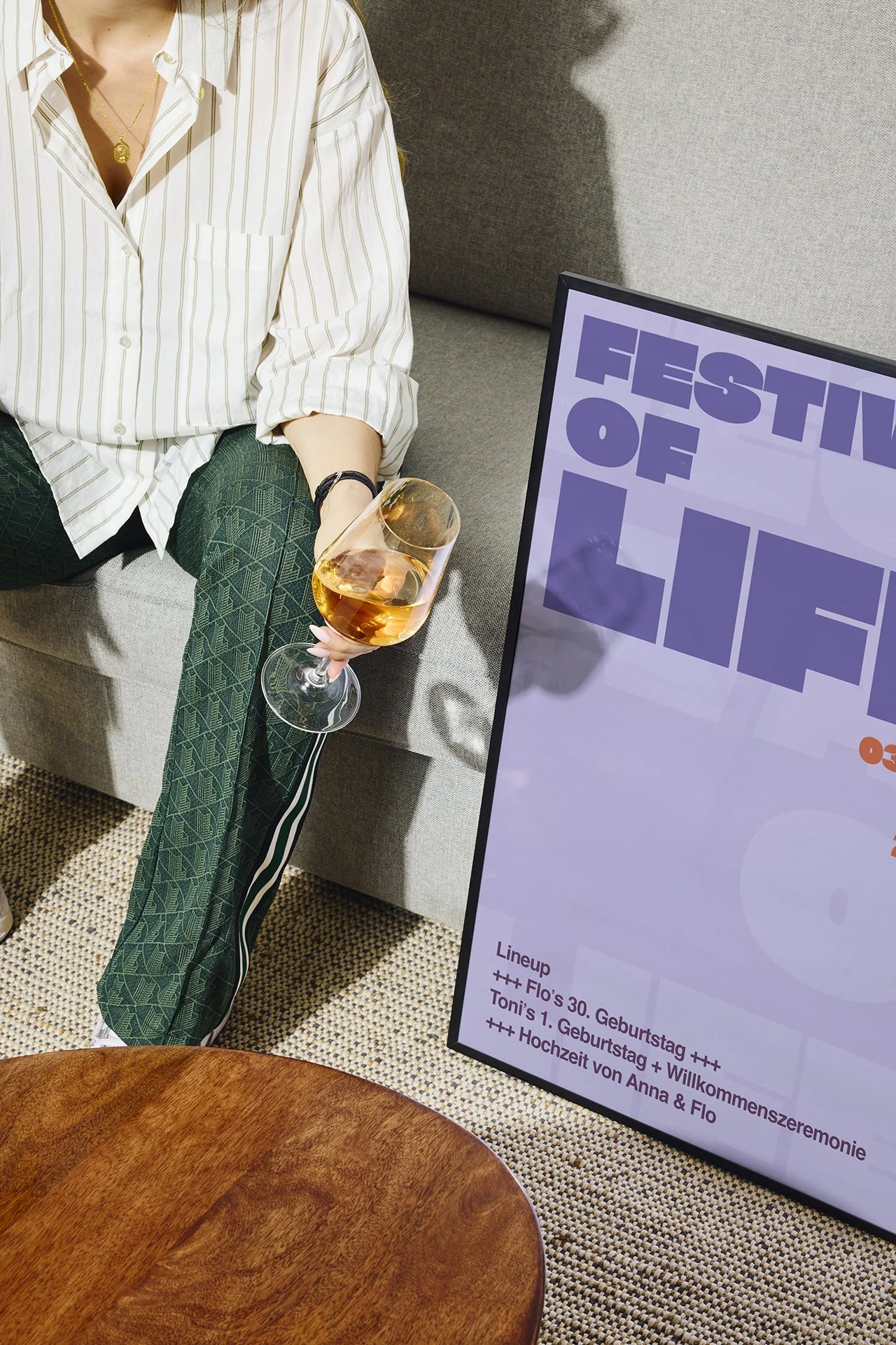 Eine Person sitzt auf einem Sofa, hält ein Glas Roséwein, trägt eine weiße Hemd und grüne Sportswear, neben einem Plakat mit Veranstaltungstext.