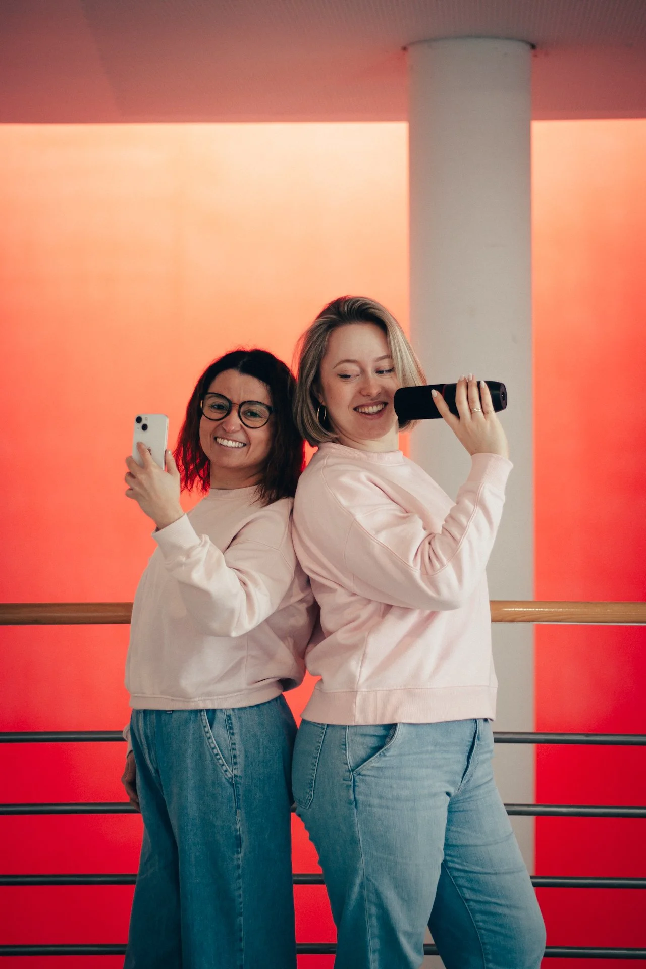 Zwei Frauen posieren für ein Selfie in einem Raum mit rotem Hintergrund, sie tragen rosa Sweatshirts und Jeans, eine lächelt in die Kamera, die andere blickt auf ihr Smartphone.