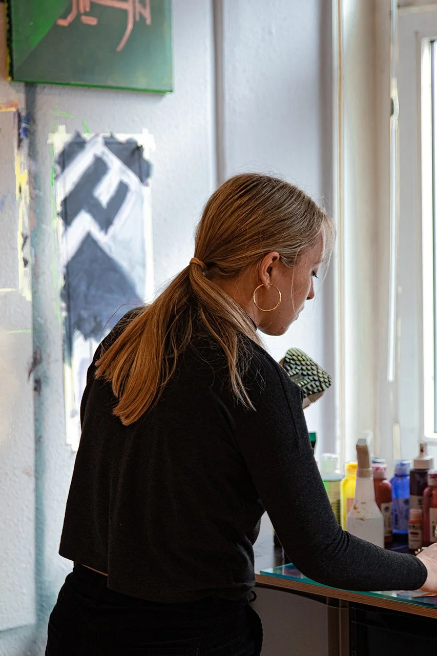Eine Frau mit langen, hellbraunen Haaren und einem schwarzen Pullover steht an einem Tisch mit bunten Acrylfarben und Pinseln vor einem Fenster. Sie ist in einer kreativen Umgebung tätig.