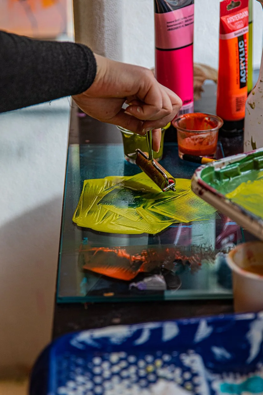 Hand, die eine Glasscheibe mit gelber Farbe bemalt, auf einem Tisch mit Pinsel und Farbtuben.