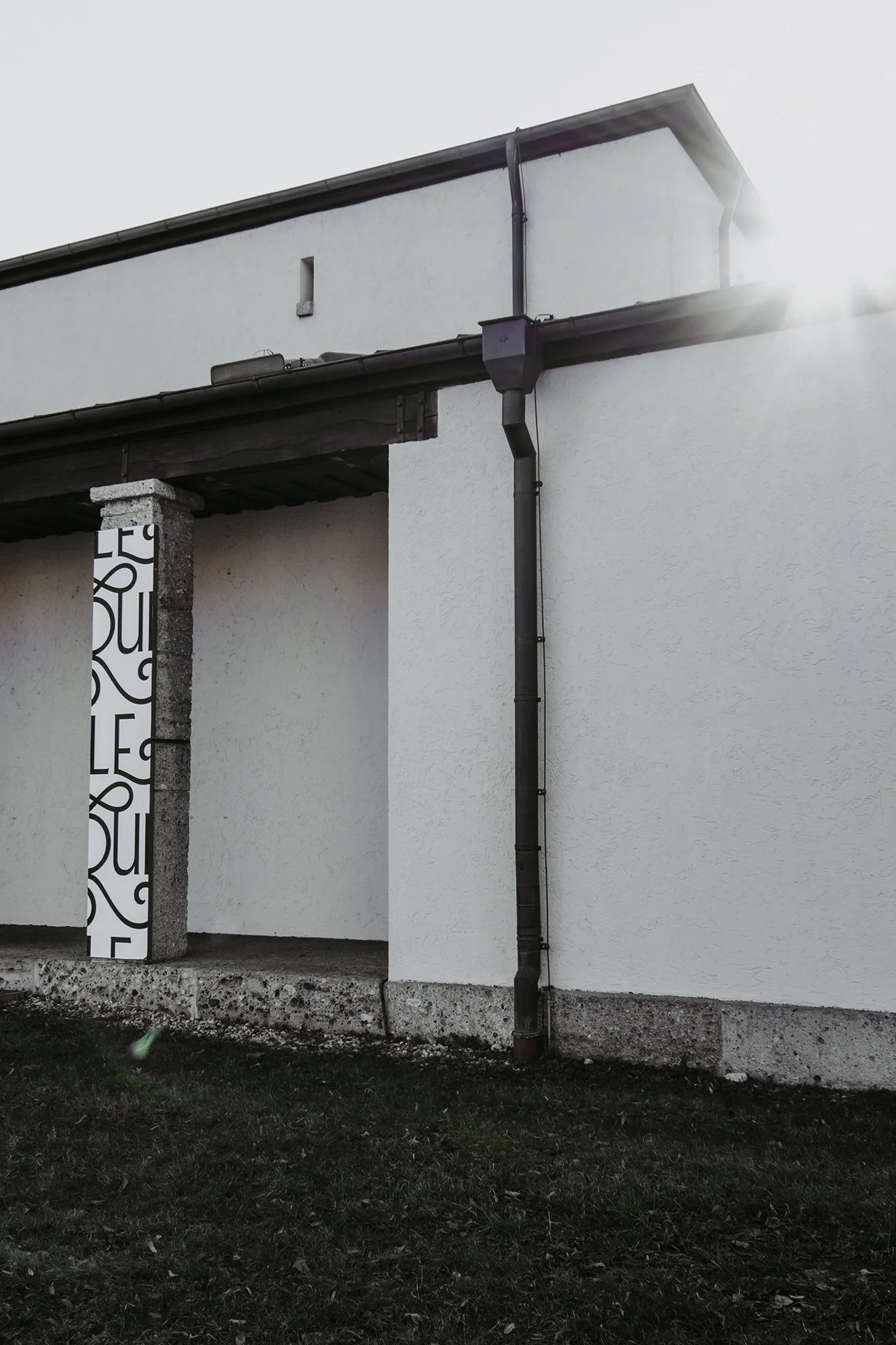 Ansicht eines weißen Hauswand mit Dachrinne und Fallrohr, im Vordergrund ein Rasen. Am linken Rand eine Betonsäule mit schwarzer und weißer Mustertapete.
