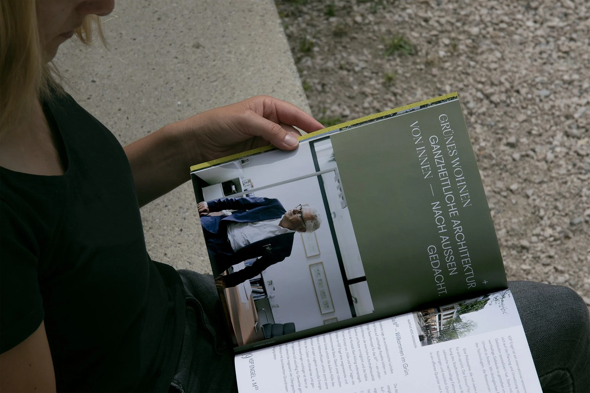 Eine Person liest ein Buch im Freien, sitzend auf einer Bank. Das Buch zeigt ein Bild eines alten Mannes in einem schwarzen Anzug und einem weißen Hemd, der in einem modernen, hell erleuchteten Raum steht.