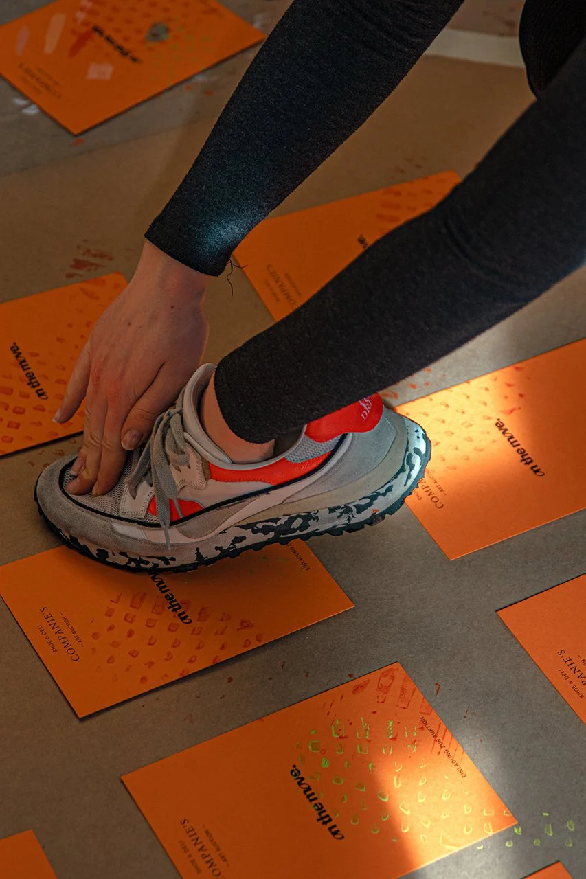 Person in schwarzer Sportkleidung, die einen grauen und orange Sportschuh auf orangen Informationskarten stempelt.