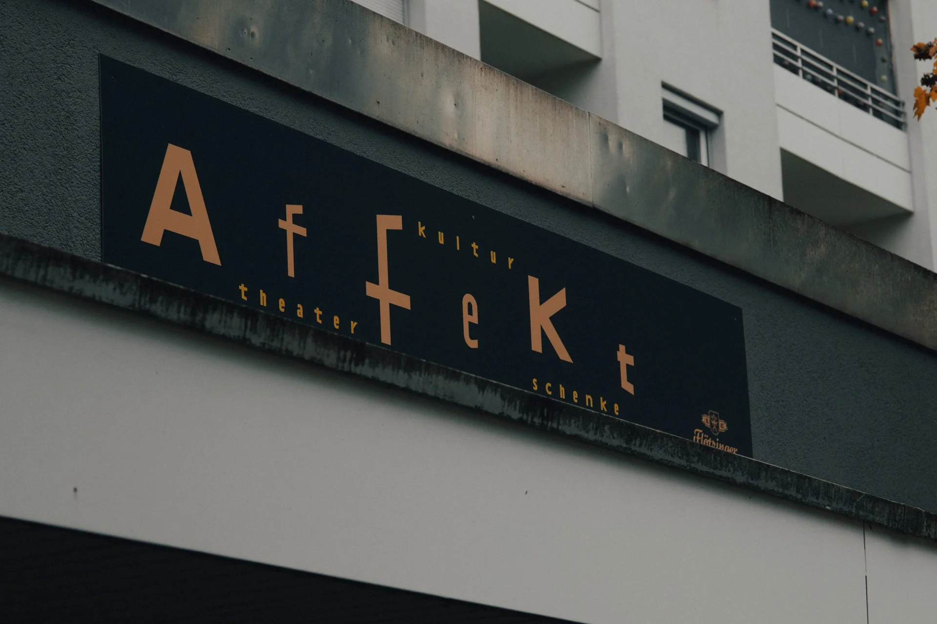 Das Bild zeigt ein schwarzes Schild mit dem Schriftzug 'Aftekt kultur theather schenke' an einem Außenwand eines Gebäudes, darüber mehrere Etagen mit Balkonen und Fenstern, und eine Herbstblattverzierung am oberen rechten Rand.