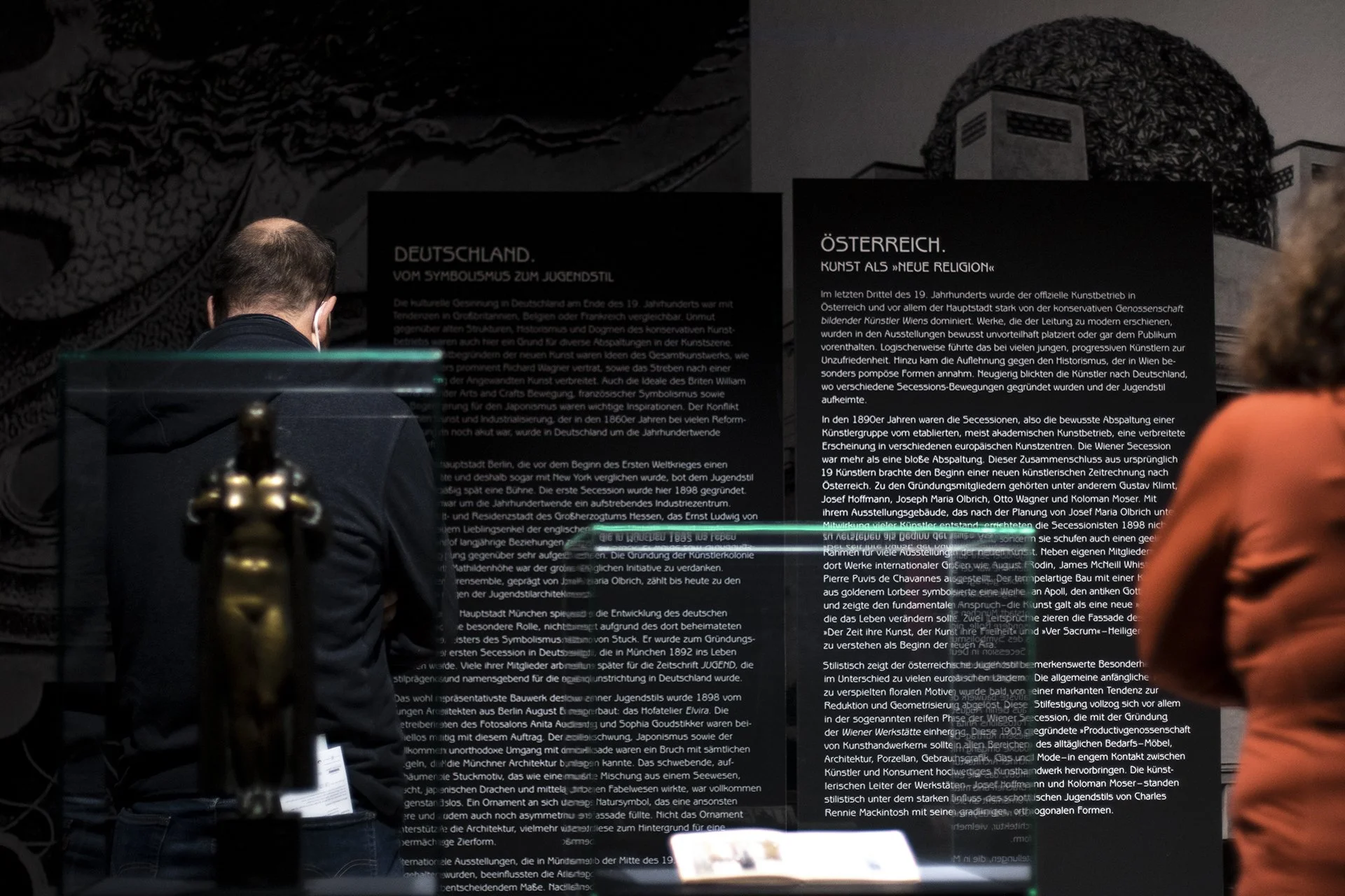Eine Person mit dunkler Kleidung, die eine Wand mit Text auf Deutsch betrachtet, wahrscheinlich in einer Ausstellung oder einem Museum.