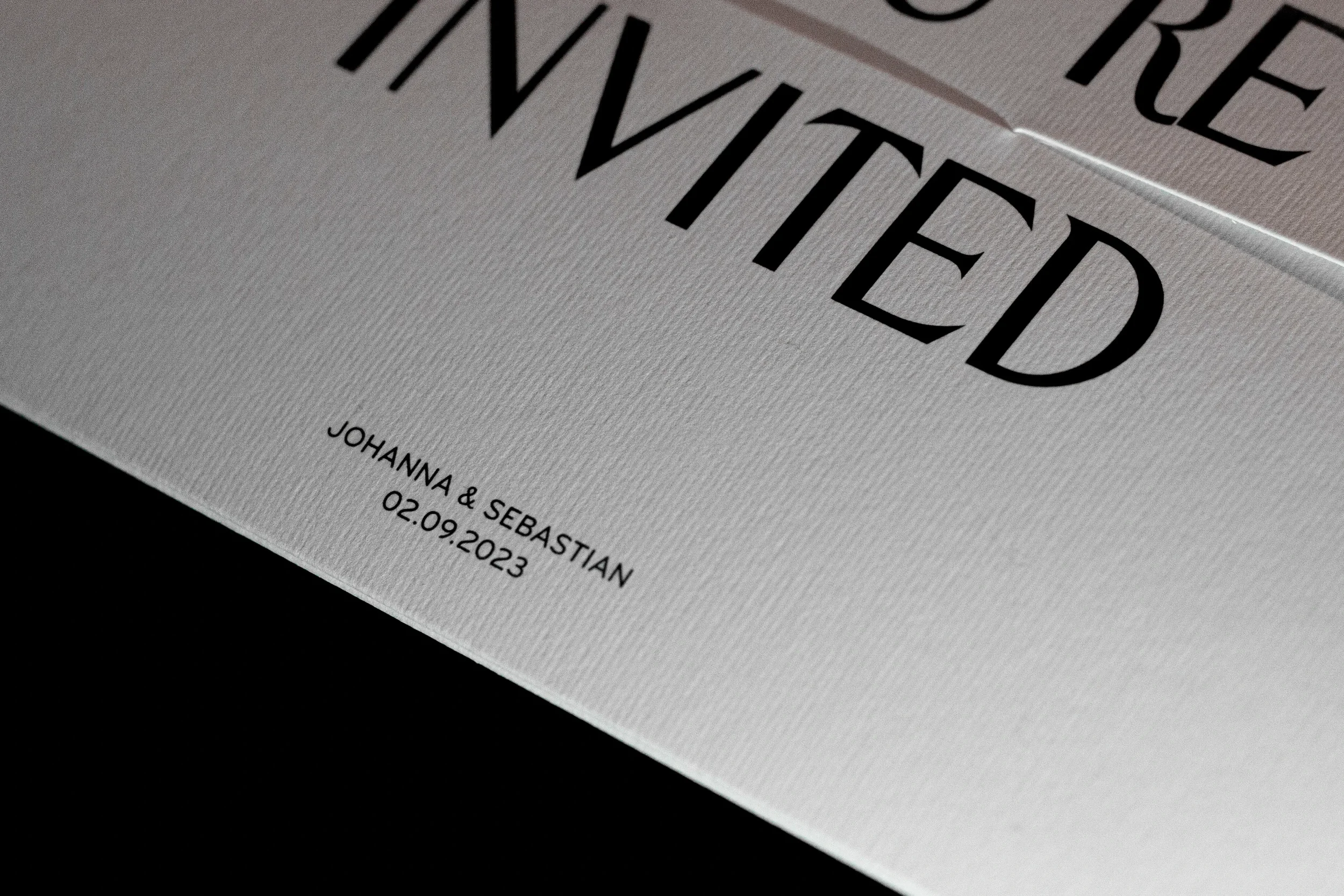 Nahaufnahme einer weißen Einladungskarte mit schwarzer Schrift, die den Namen "INVITED" sowie die Namen "Johanna & Sebastian" und ein Datum "02.09.2023" zeigt.