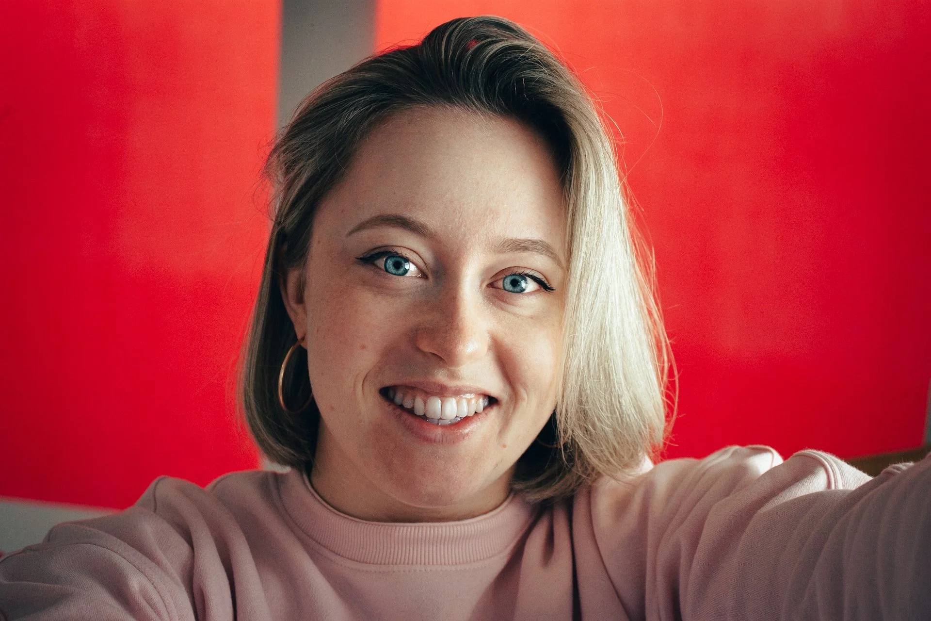 Junge Frau mit blonden Haaren, blaue Augen und großen Ohrringen, lächelnd in die Kamera fotografiert, vor einem pinken Hintergrund.
