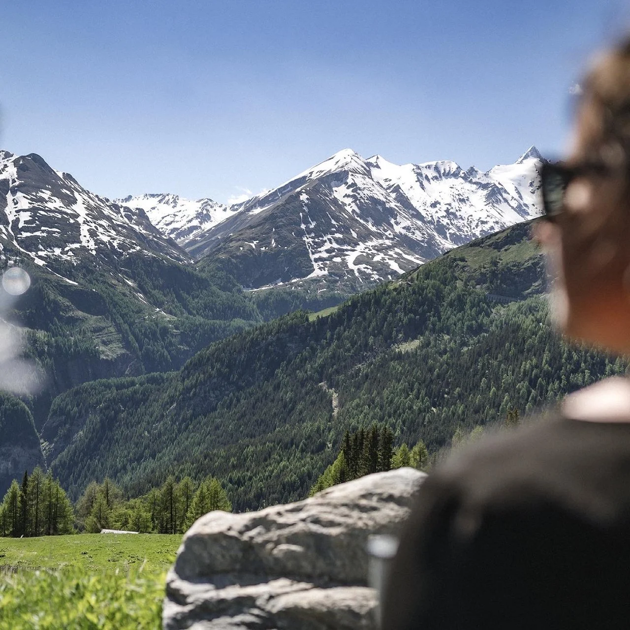 Eine Person sitzt auf einem Stein mit Blick auf grüne Hügel und schneebedeckte Berge im Hintergrund.