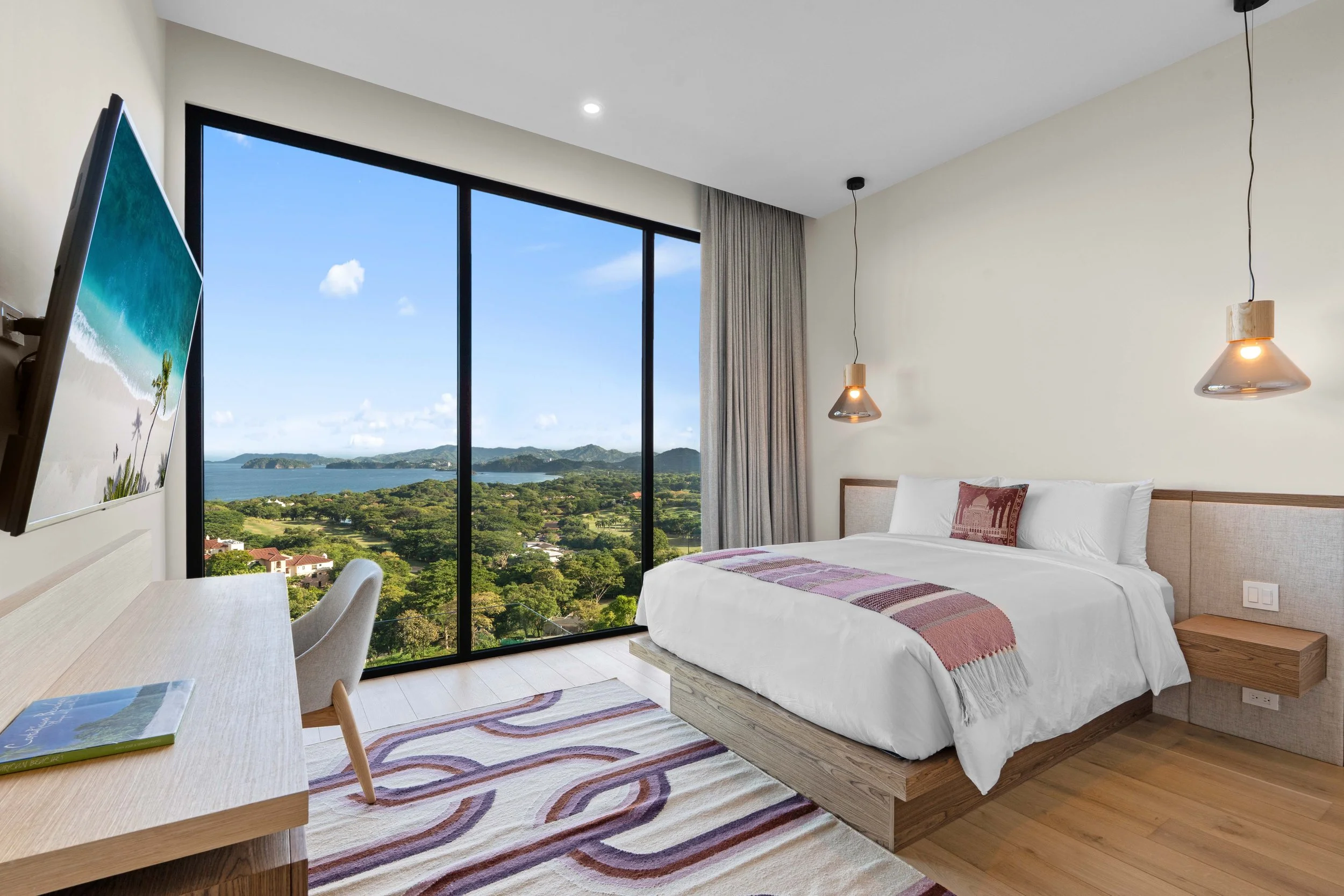 Modern hotel room with a large window showing a scenic view of greenery, water, and distant islands, a bed with white linens, a wooden headboard, hanging pendant lights, a rug, a desk with a book, and a wall-mounted television.