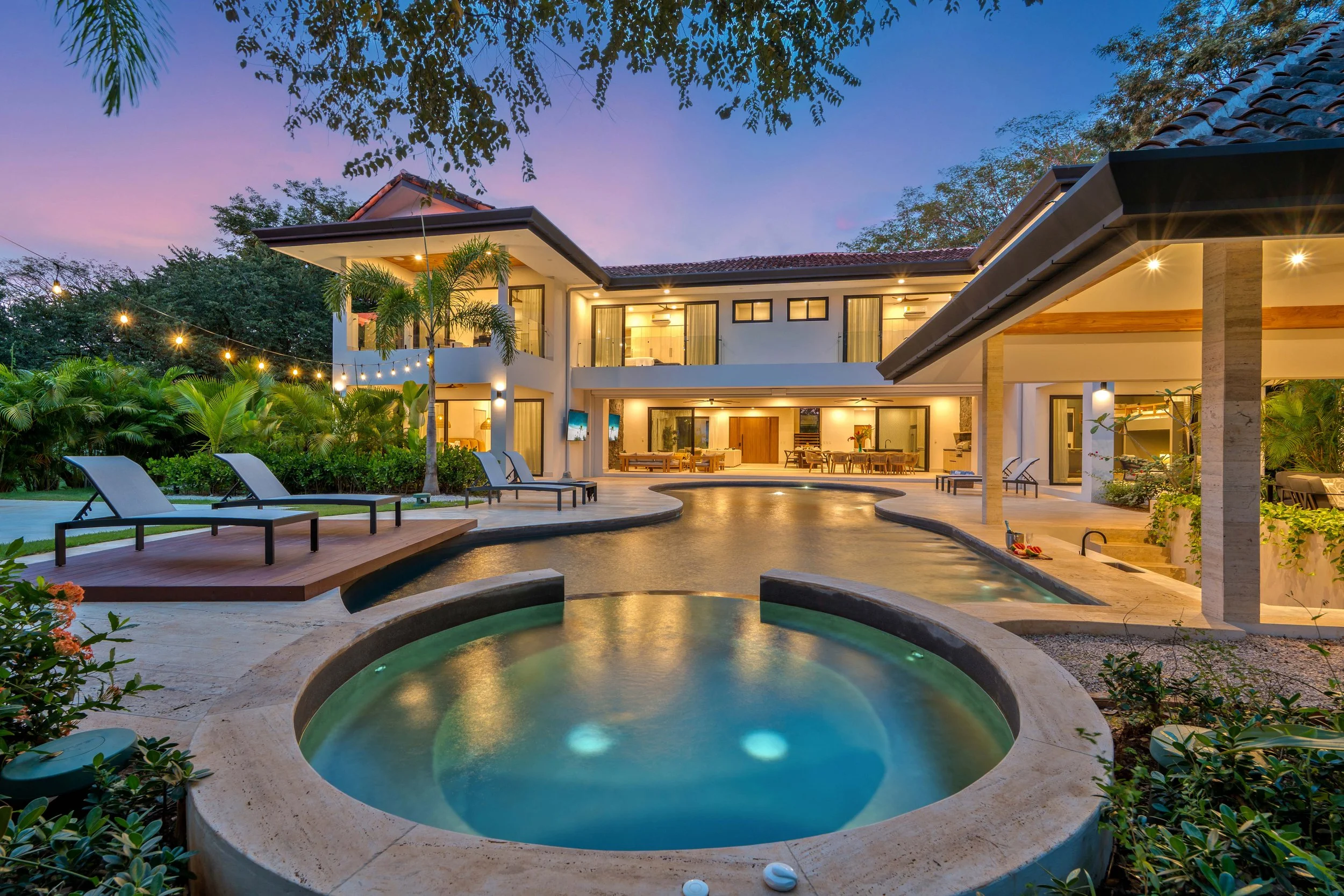 Luxury modern house with pool area at sunset, illuminated outdoor lighting, lounge chairs by the pool, tropical plants, and a covered patio.