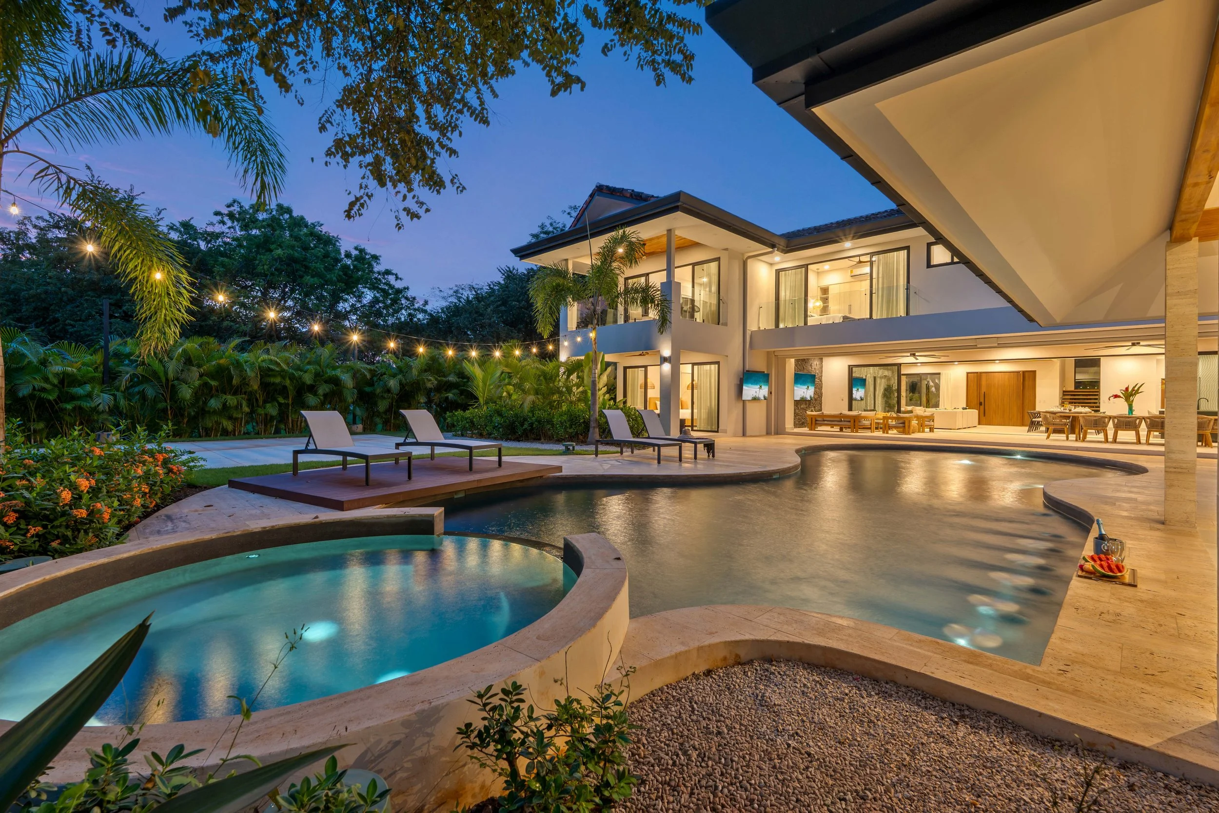 A modern house in Tamarindo, Costa Rica with a pool and hot tub, outdoor lounge chairs, lush greenery, and string lights at dusk.