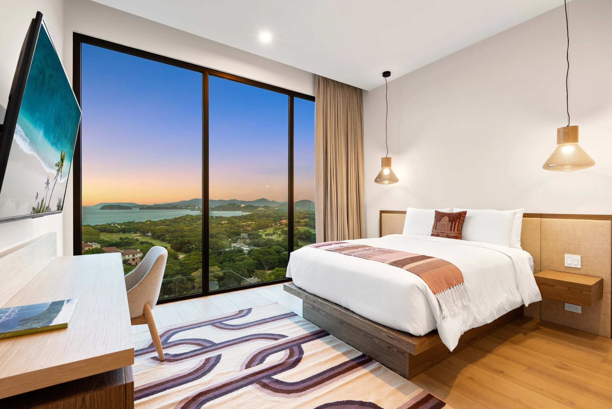 Modern hotel room with a large bed, hanging pendant lights, a view of a sunset over a landscape with water, a desk with a chair, and a television.