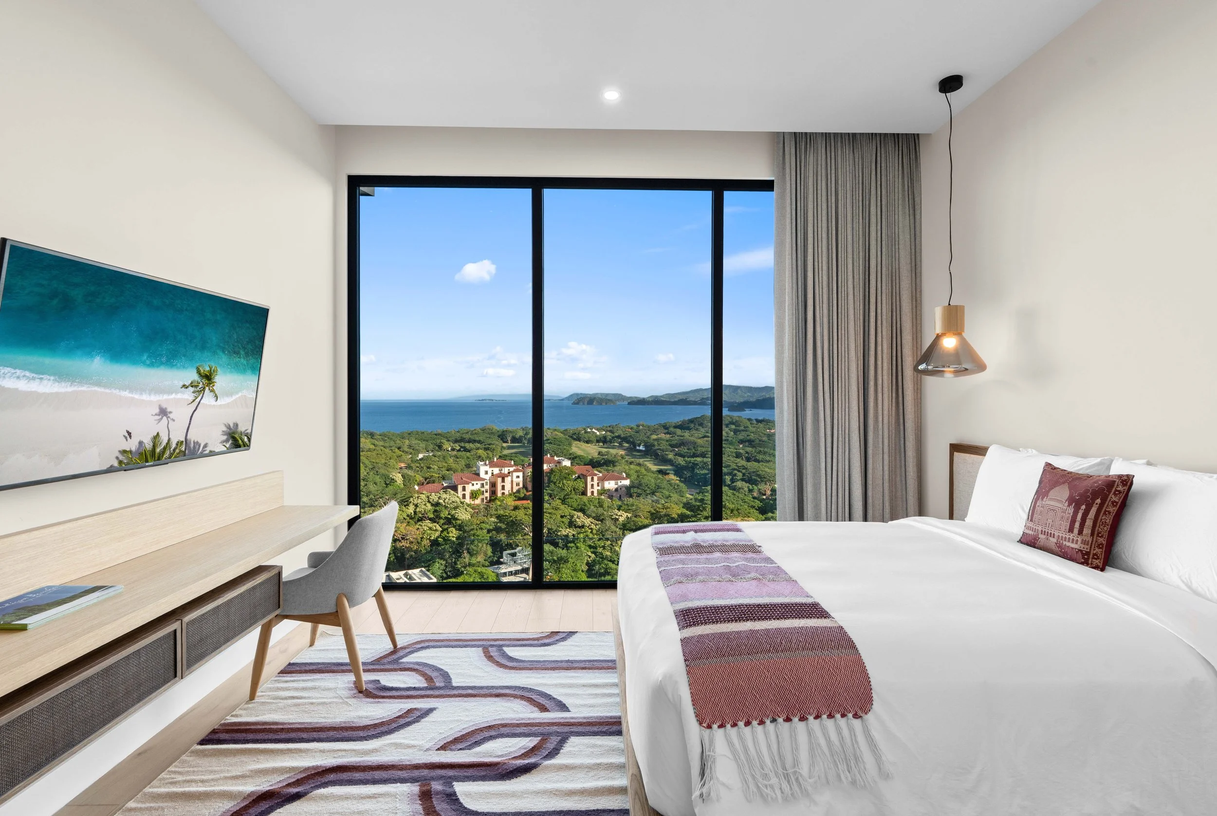 Modern hotel room with a large window showing a scenic view of a green landscape, water, and blue sky, featuring a bed with white linens and a decorative pillow, a wall-mounted TV, a desk with a chair, and a hanging lamp.