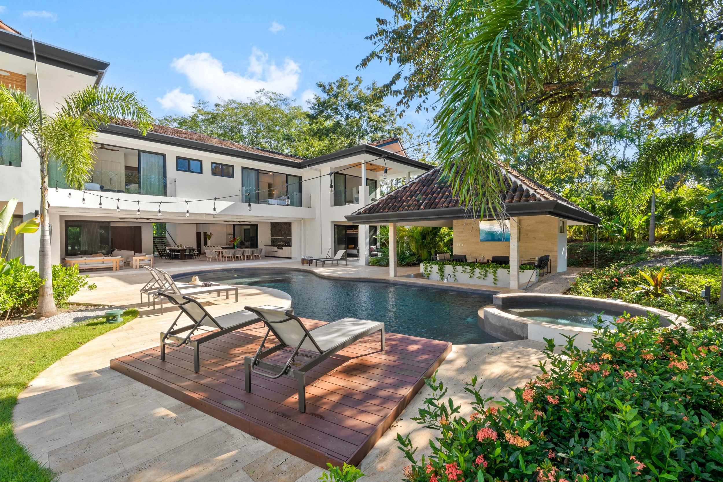 Luxury house with a swimming pool, lounge chairs, hot tub, and tropical landscaping under a partly cloudy sky.