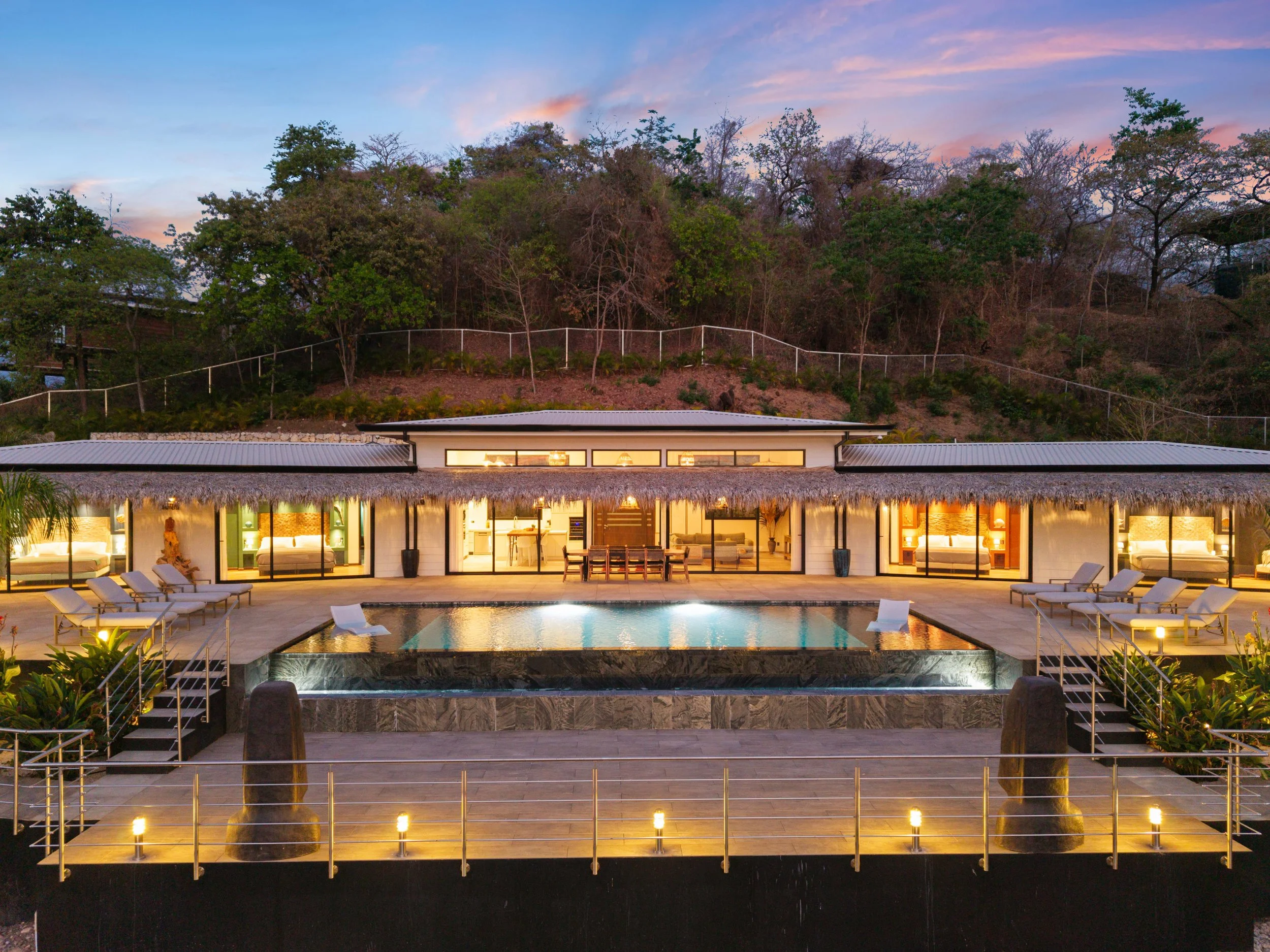 Luxurious modern house in Tamarindo, Costa Rica with floor-to-ceiling windows, a swimming pool, and outdoor lounge chairs, set against a hillside with trees at dusk.