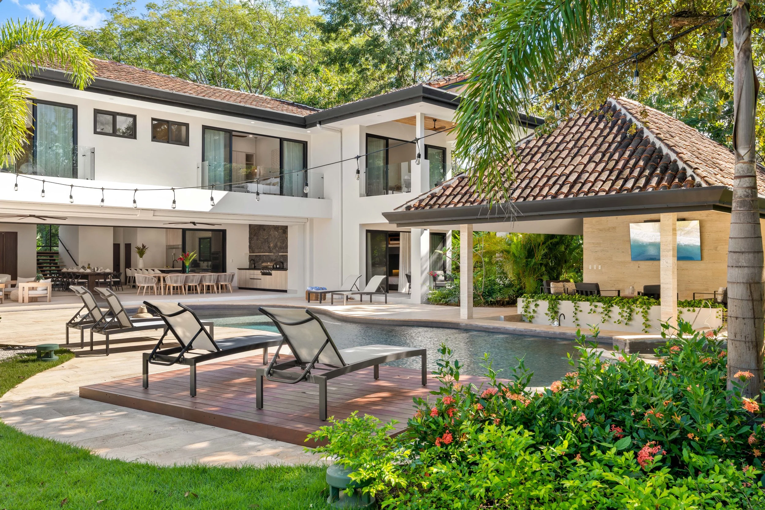 Luxury modern house with swimming pool, outdoor seating, lounge chairs, lush greenery, and a shaded patio area.
