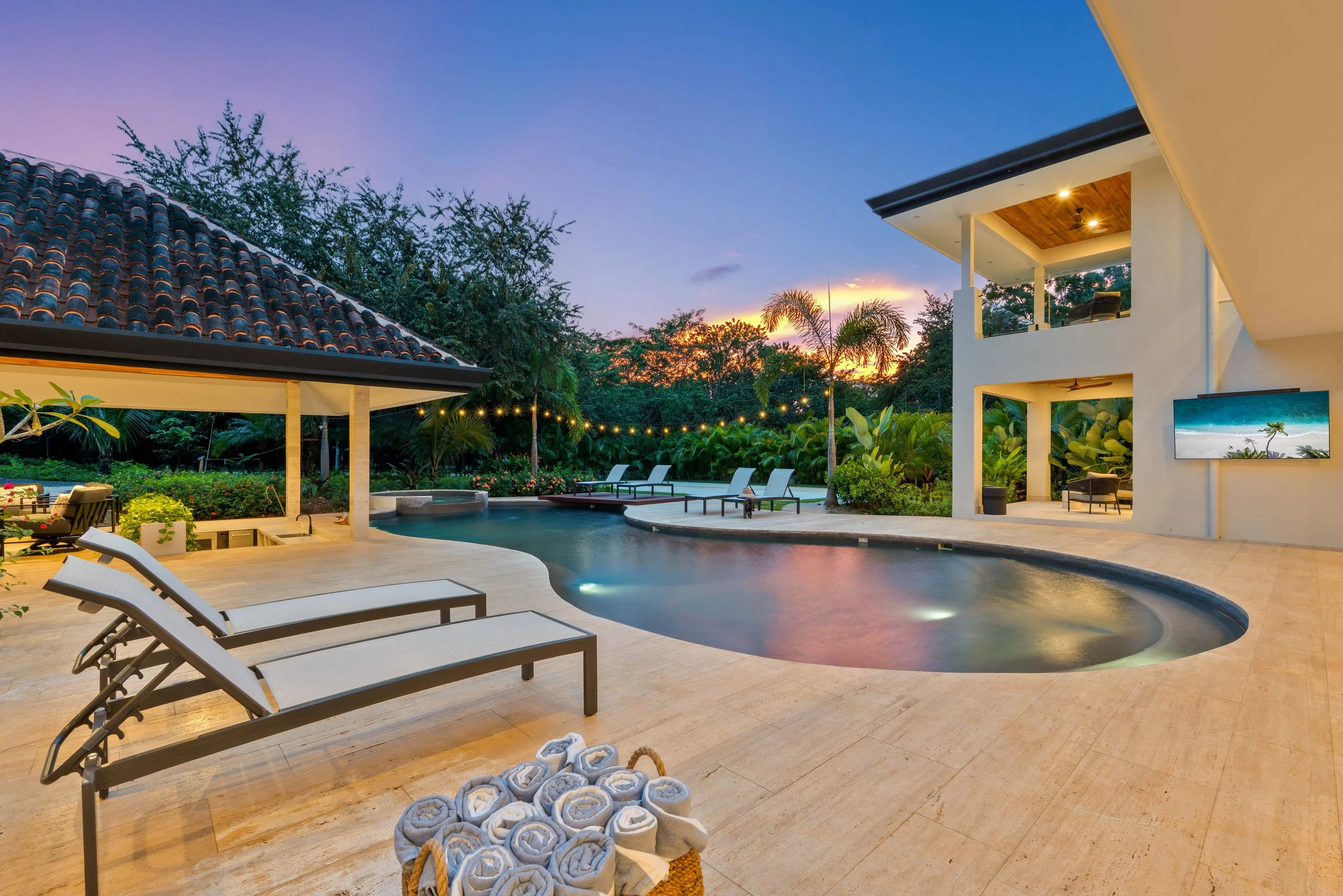 Luxury backyard with a swimming pool, loungers, outdoor seating, string lights, lush greenery, and a sunset sky.