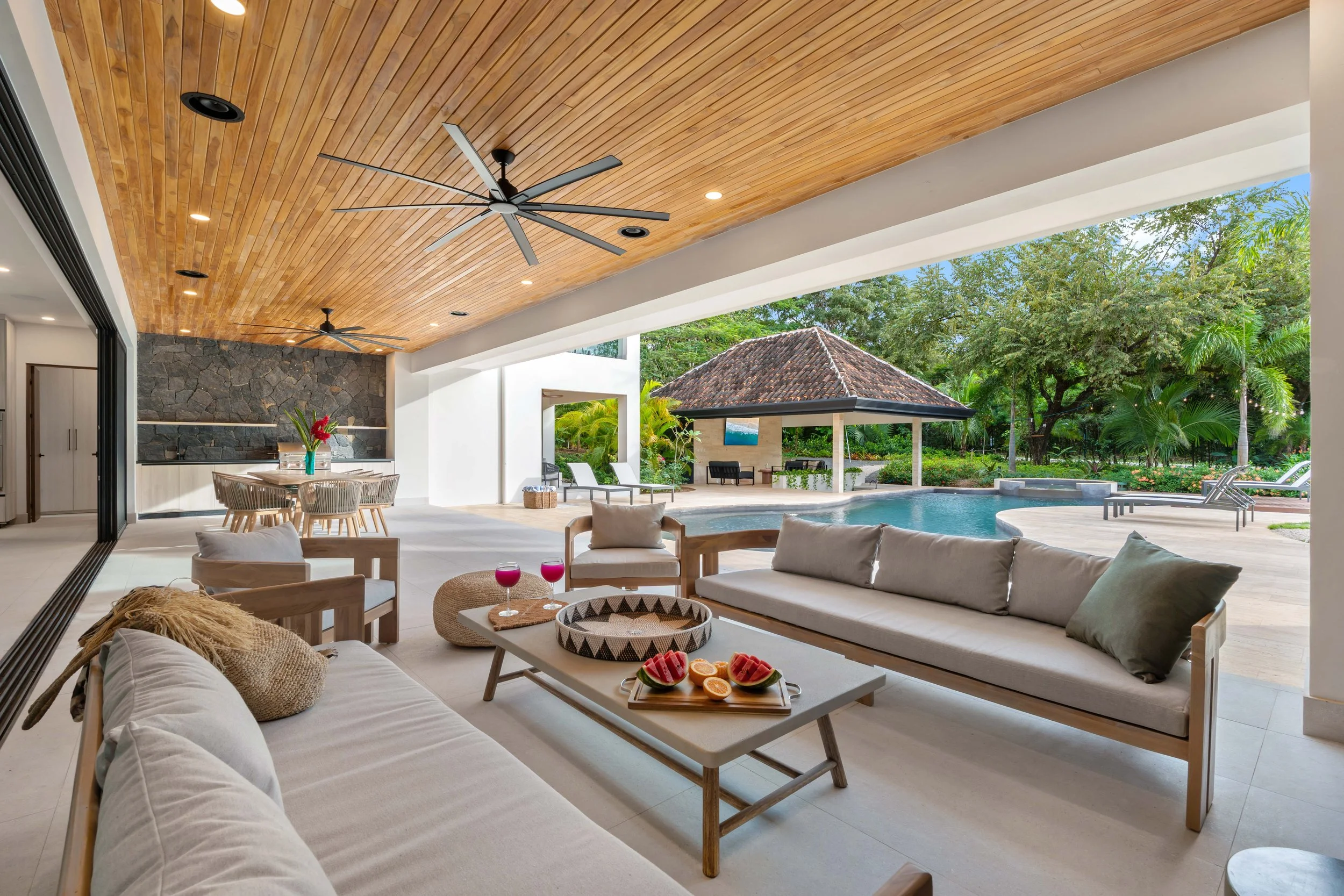 Open-air living space with beige couches, a wooden table with drinks and fruit, overlooking a pool and outdoor pavilion with trees in the background.