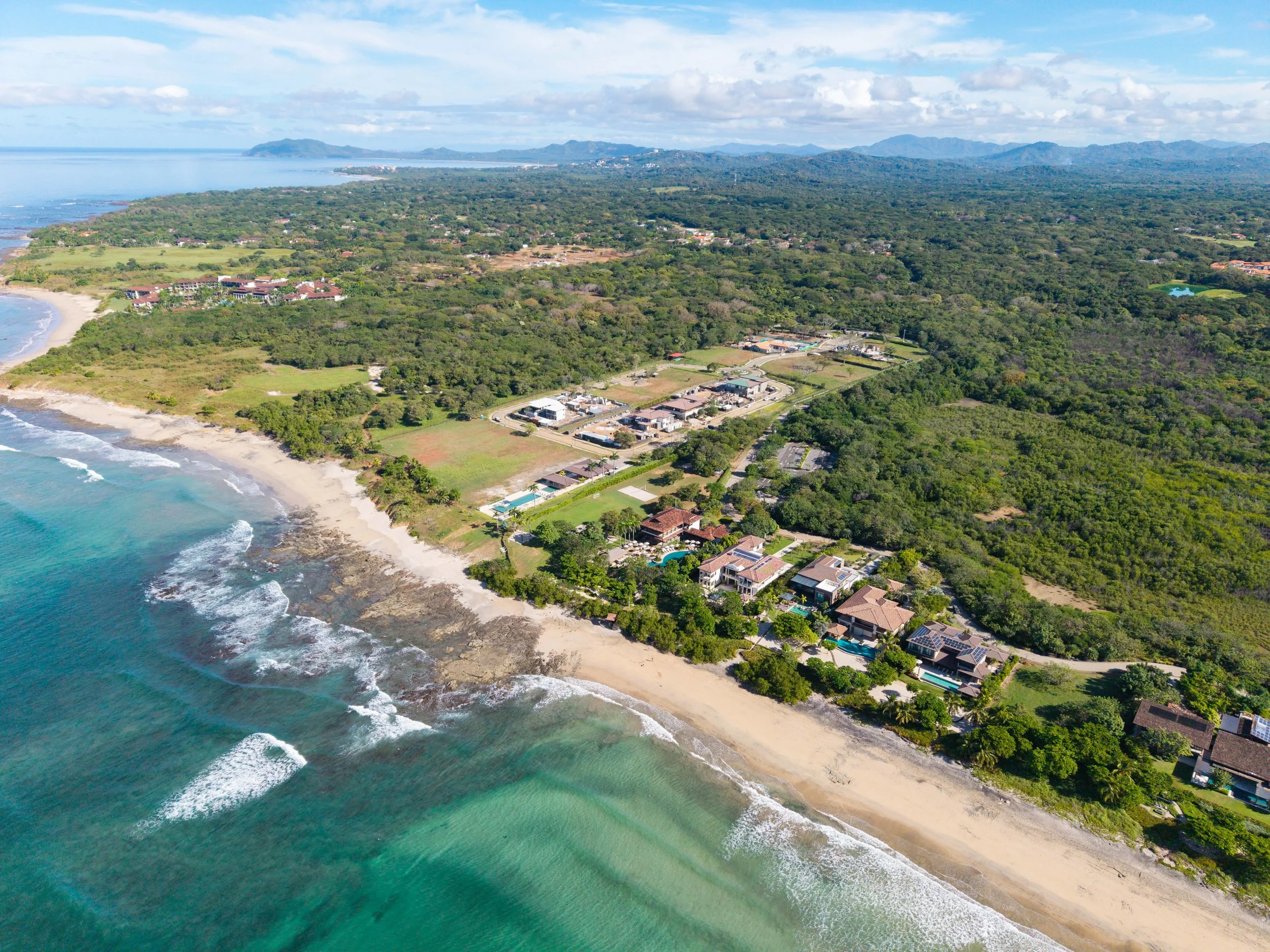 Aerial view of a coastal neighborhood with sandy beaches, turquoise ocean waters, green trees, and houses with swimming pools, backed by a lush forest and mountains in the distance.