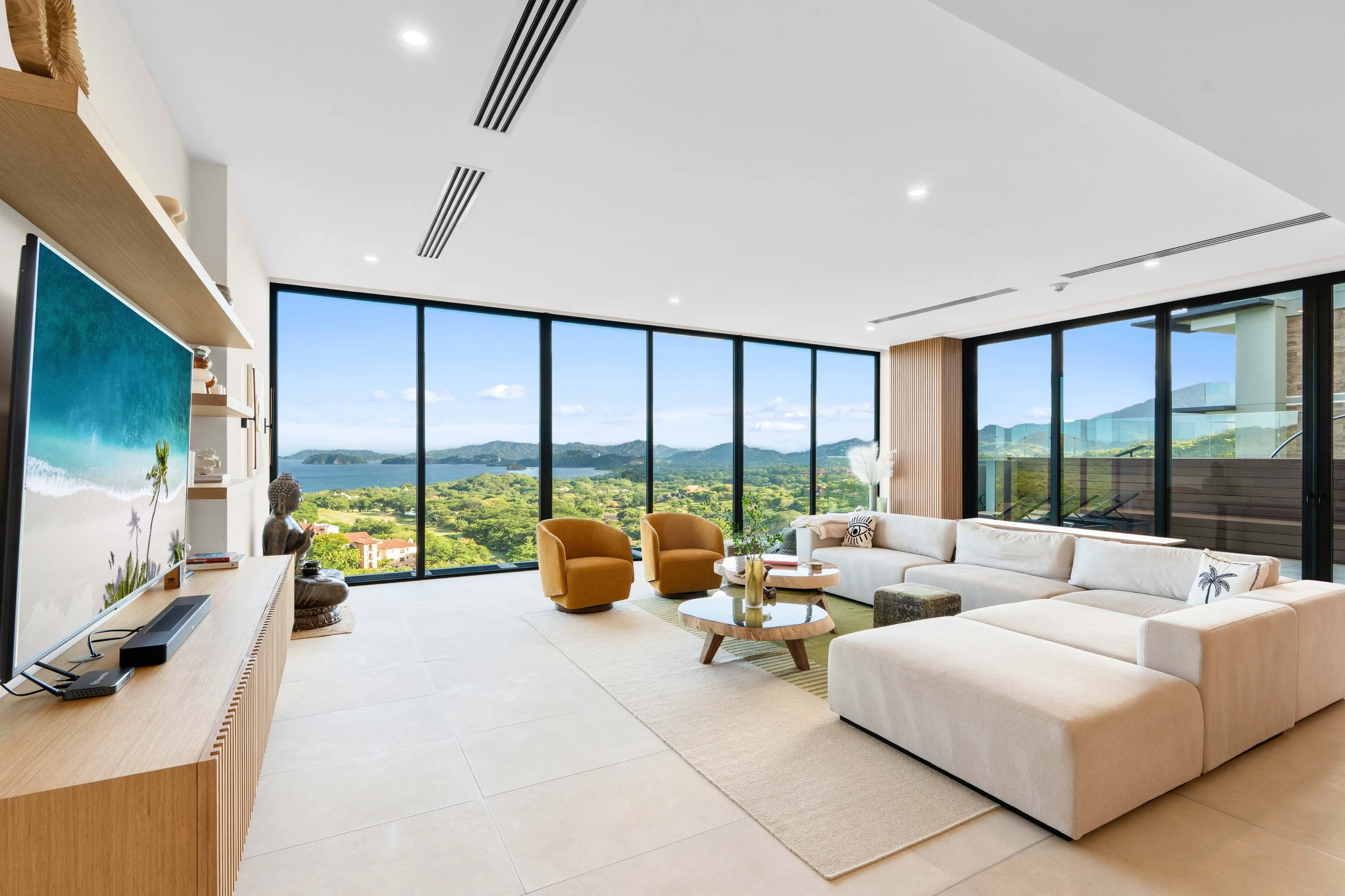 Modern living room with large floor-to-ceiling windows overlooking a green landscape, mountains, and water. Contains a beige sectional sofa, two orange armchairs, a round coffee table, a TV with a wooden console, and decorative items.