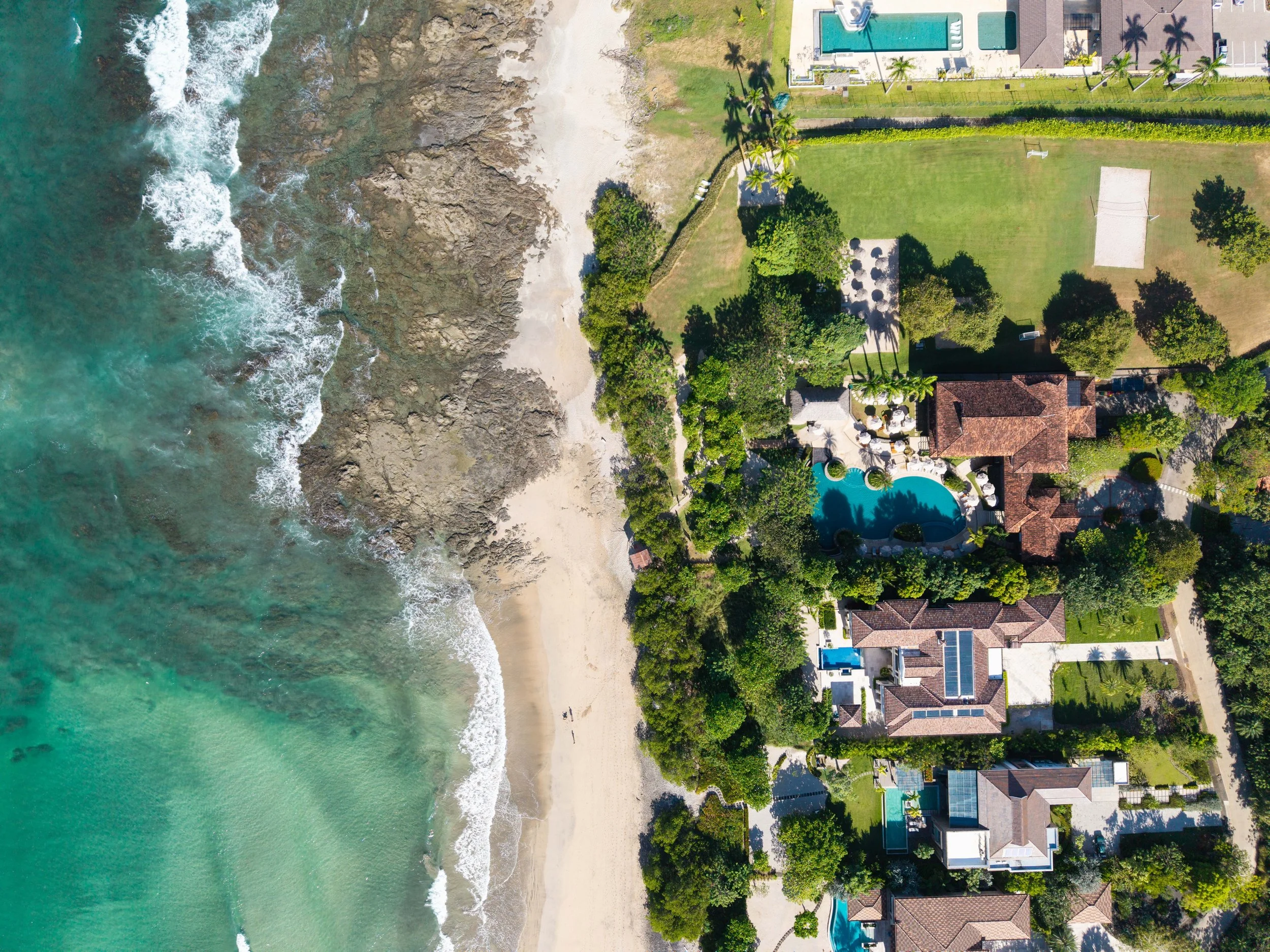 Aerial view of a beachside residential area with houses, a swimming pool, a tennis court, and a grassy yard, bordered by the ocean with waves crashing onto the sandy shoreline.