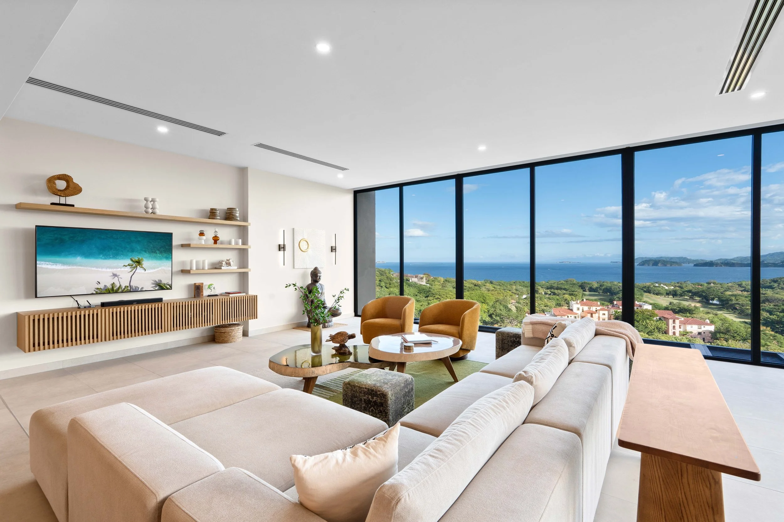 Modern living room with large floor-to-ceiling windows offering ocean and landscape view, beige sectional sofa, two orange armchairs, round wooden coffee table, TV unit, decorative shelves and sculptures, artwork, plants, and minimalist decor.
