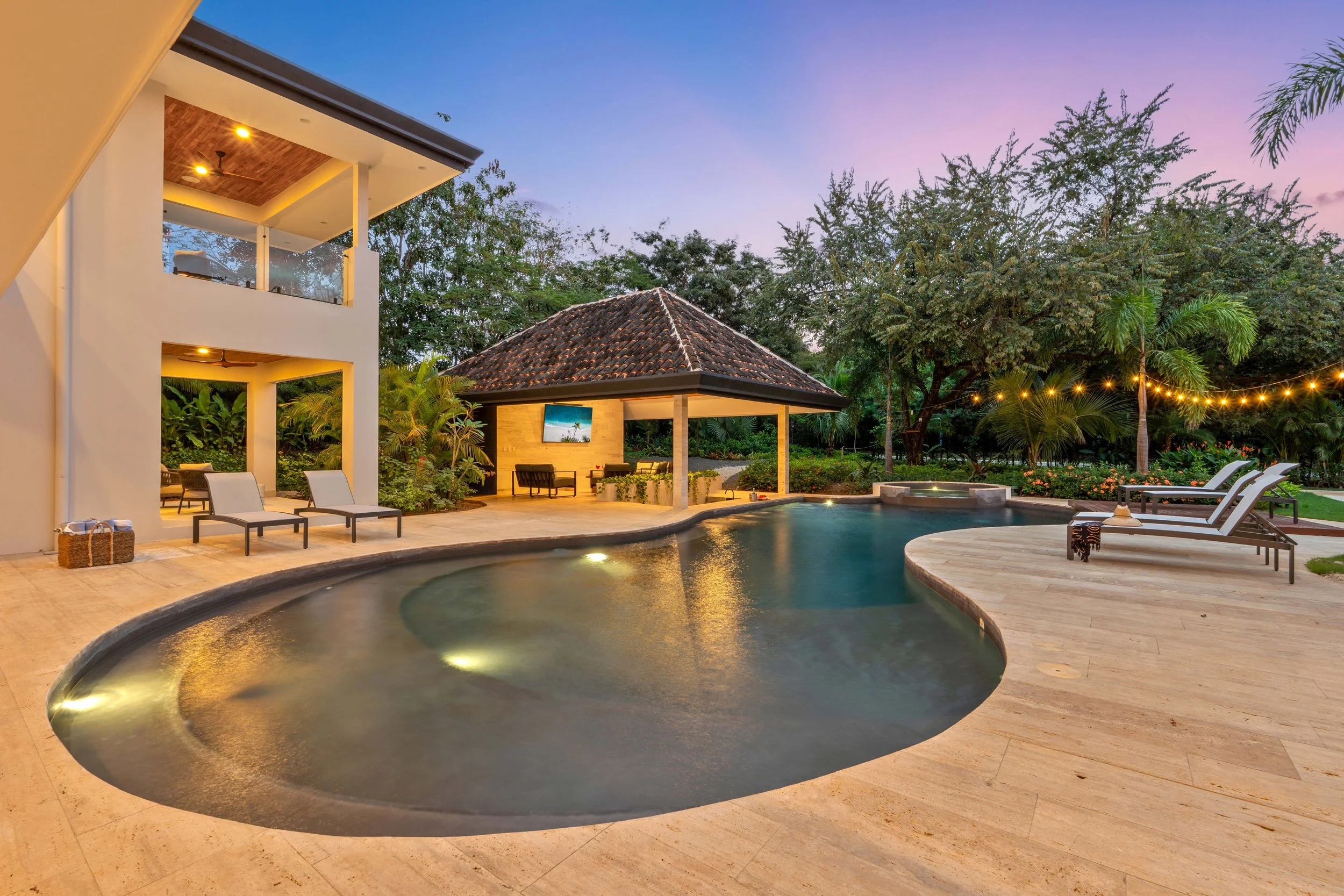 An outdoor pool area in Tamarindo, Costa Rica at sunset with lounge chairs, a gazebo, string lights, surrounded by trees and tropical plants.