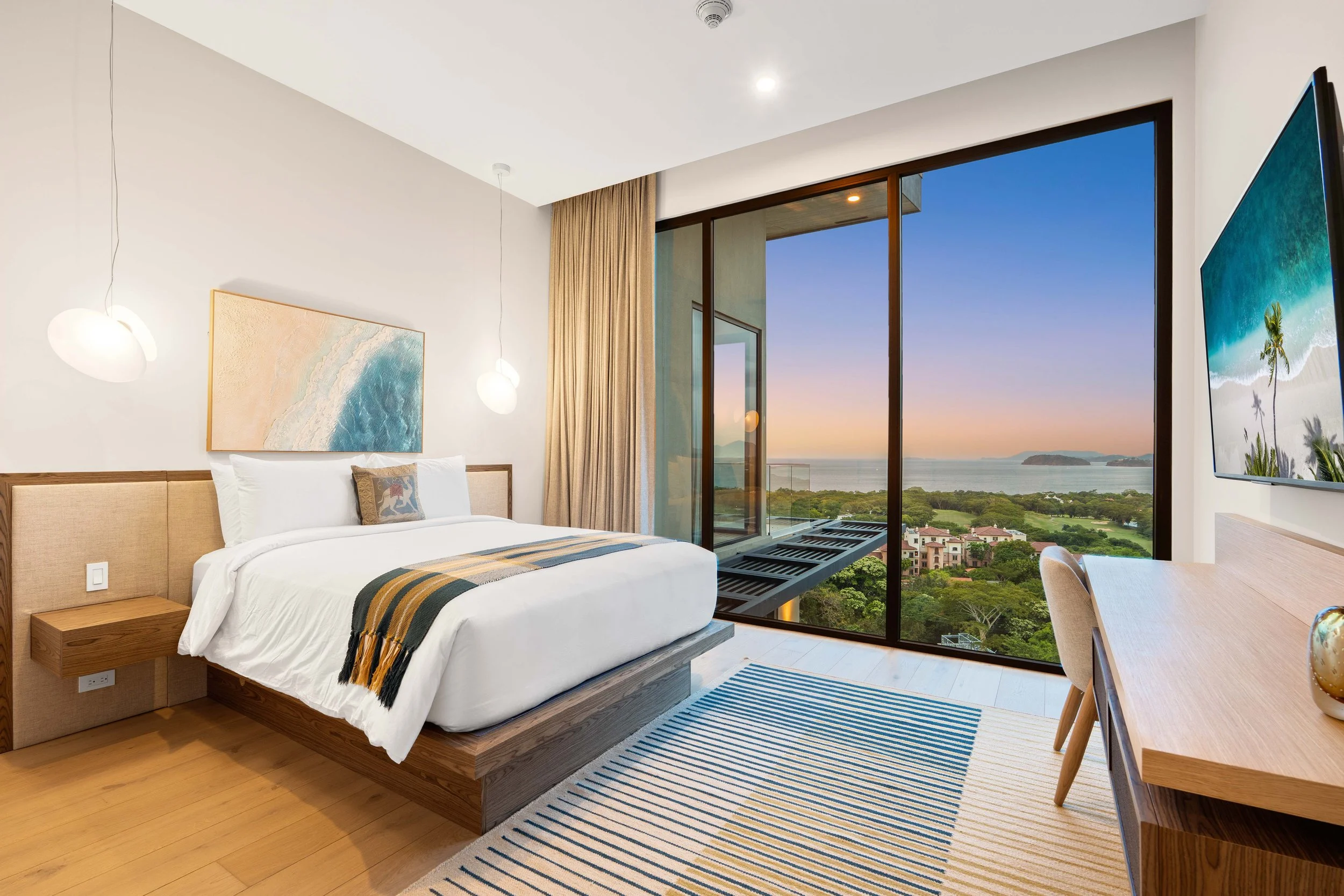 Hotel room with a large window showing a view of the ocean and greenery at sunset, modern decor, bed with white linens, and a desk.