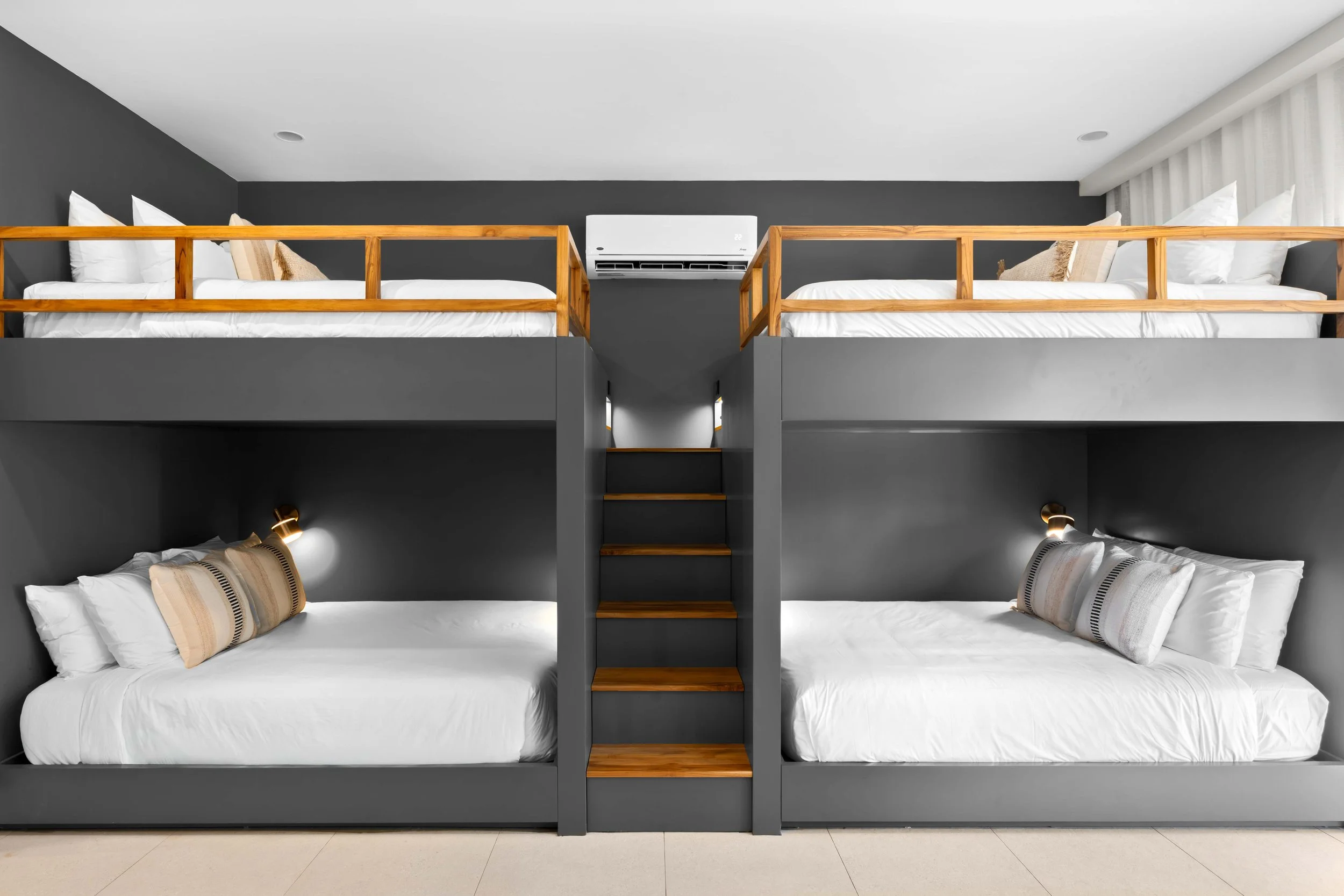 A modern bedroom with two sets of bunk beds, featuring dark gray frames, white bedding, and wooden safety railings. The room has gray walls, a ceiling-mounted air conditioning unit, and small wall-mounted reading lamps above each bed.