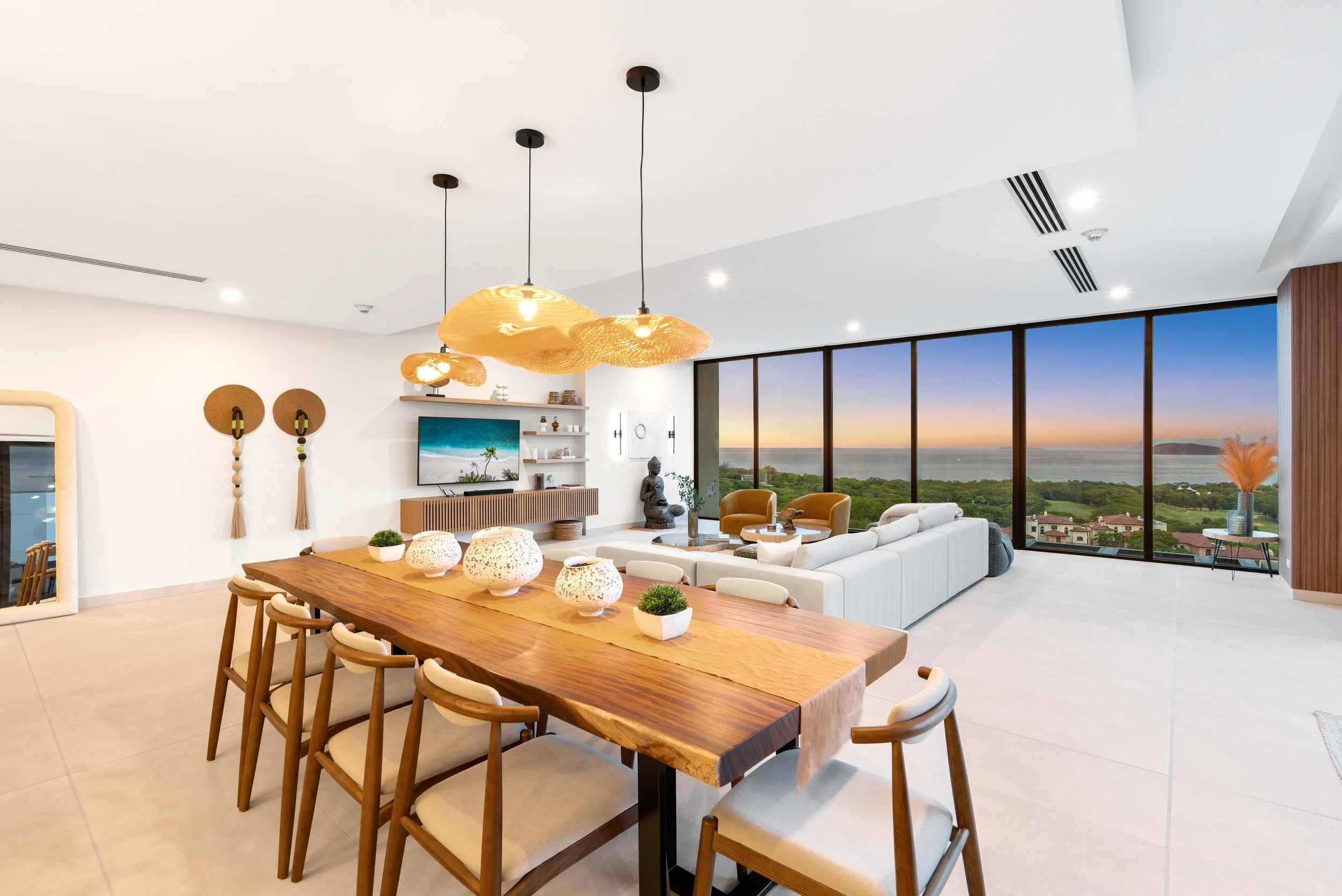 Modern living room with large windows showing a sunset over the ocean, white sectional sofa, wooden dining table with chairs, decorative vases, wall art, and hanging pendant lights.
