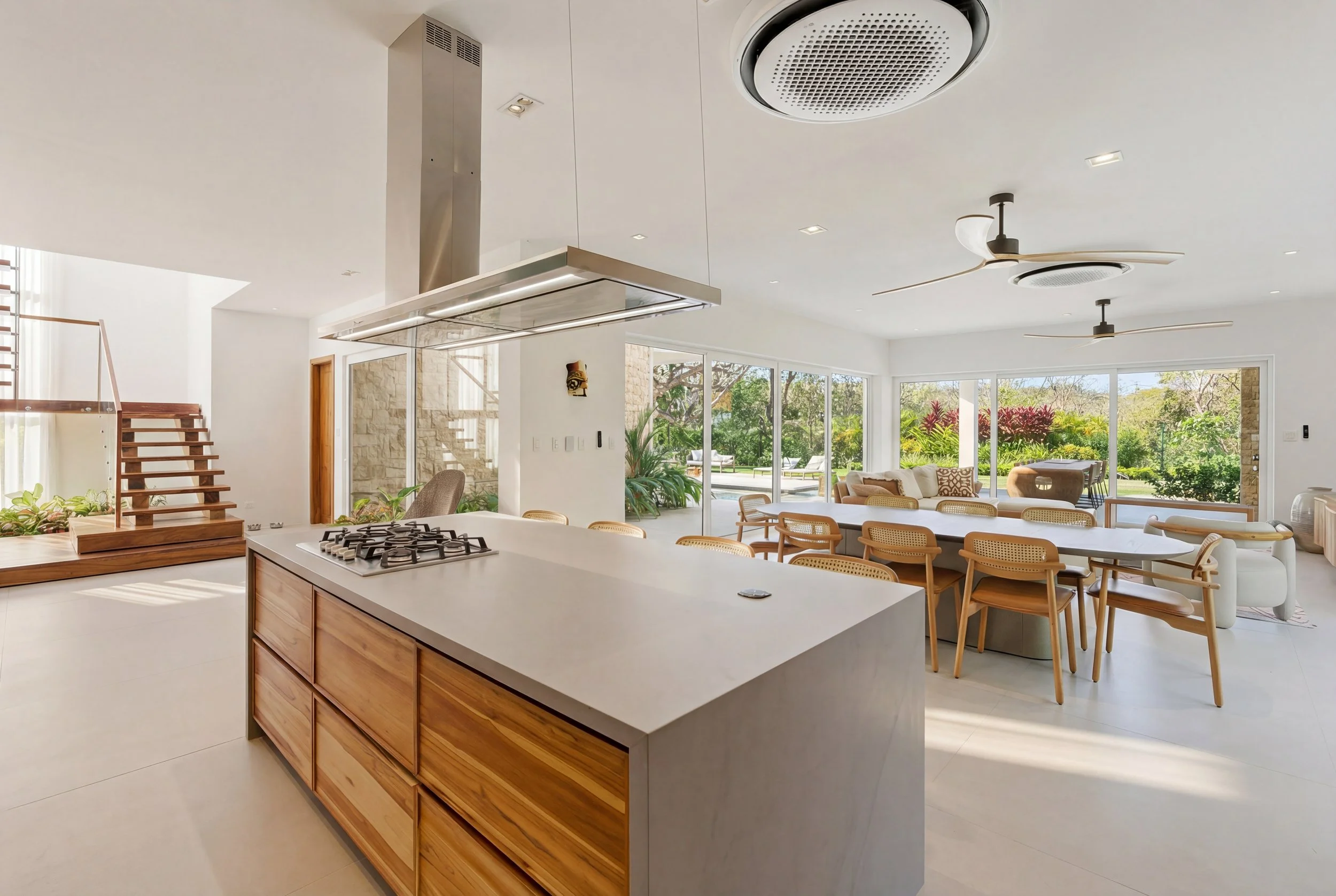 Modern open-concept kitchen and living room with large windows, wooden dining table with chairs, white sofas, ceiling fans, and a view of a lush garden outside.
