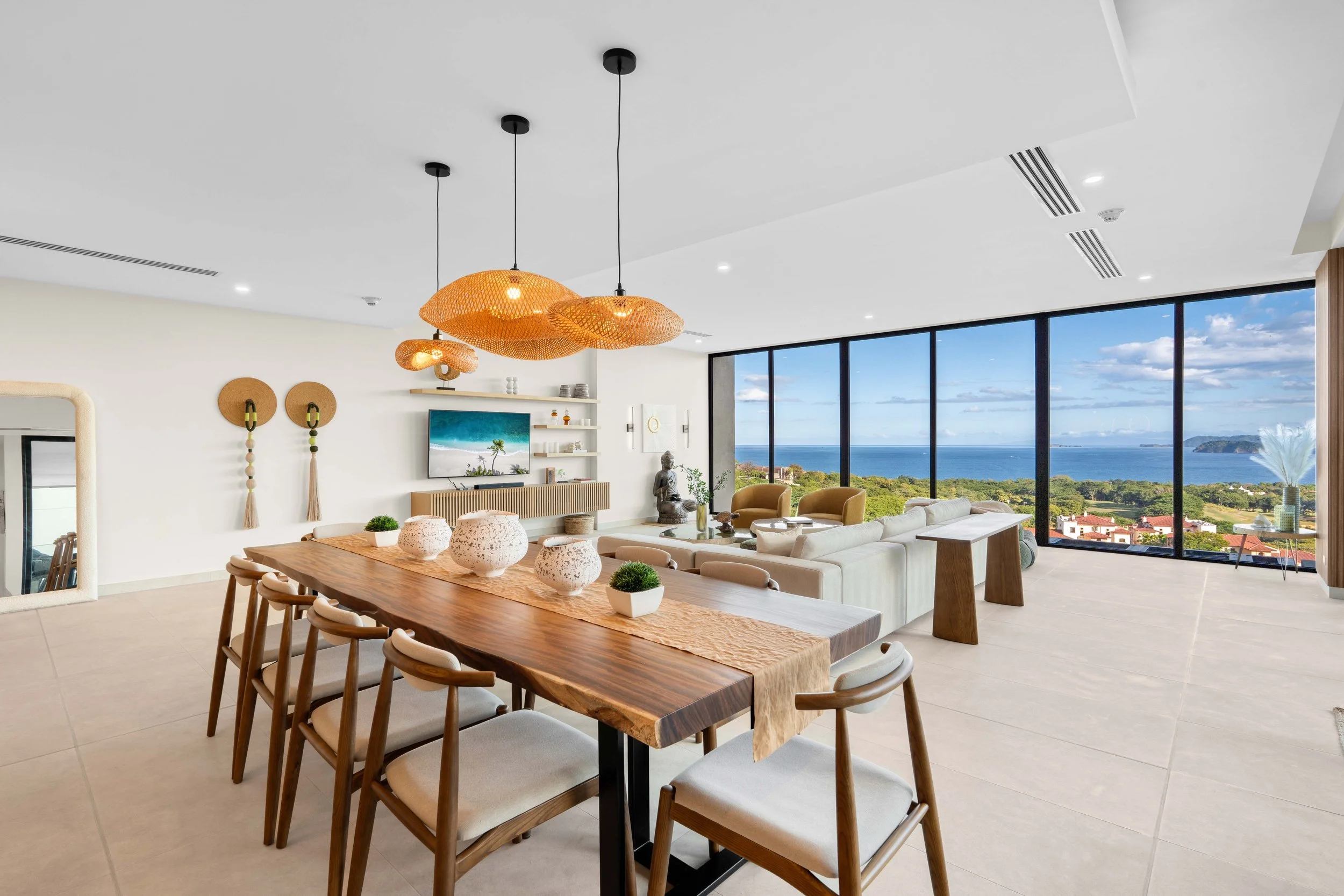 Modern living room with large windows overlooking ocean view, white sectional sofa, yellow armchairs, wooden dining table with chairs, decorative vases, pendant lights, and artwork.