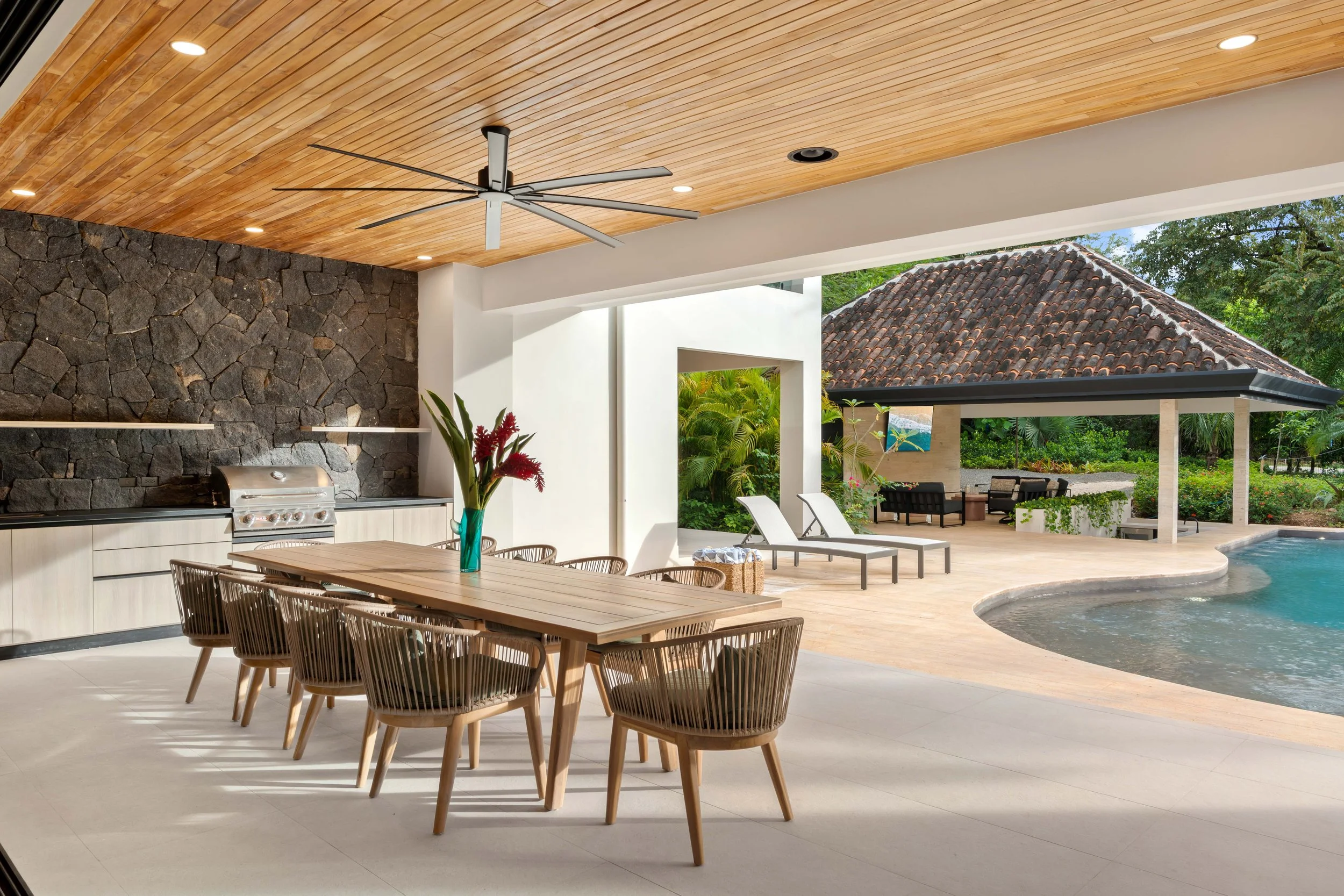Outdoor patio area with dining table, chairs, a vase with red flowers, a built-in grill, deck chairs, a swimming pool, and a covered sitting area with outdoor furniture, lush greenery, and a tiled-roof structure in the background.