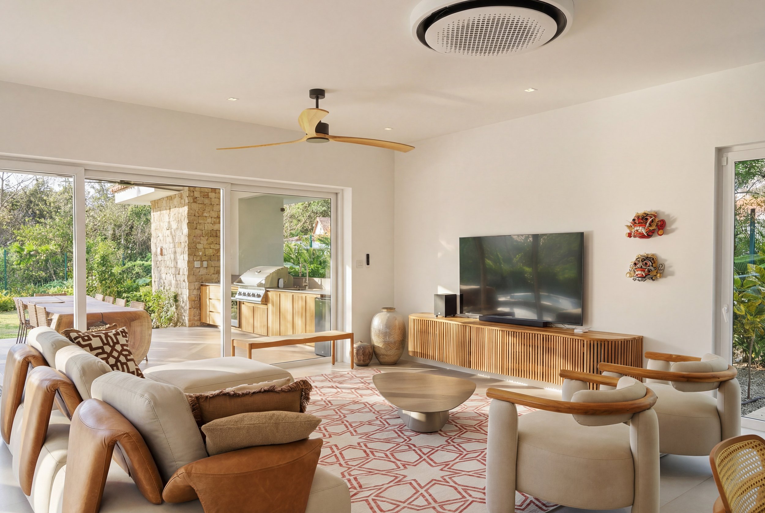 Living room with white walls, a large flat-screen TV on a wooden TV stand, two carved masks on the wall, a beige and brown sofa, two armchairs with wooden arms, a coffee table, and sliding glass doors leading to an outdoor area with a dining table an
