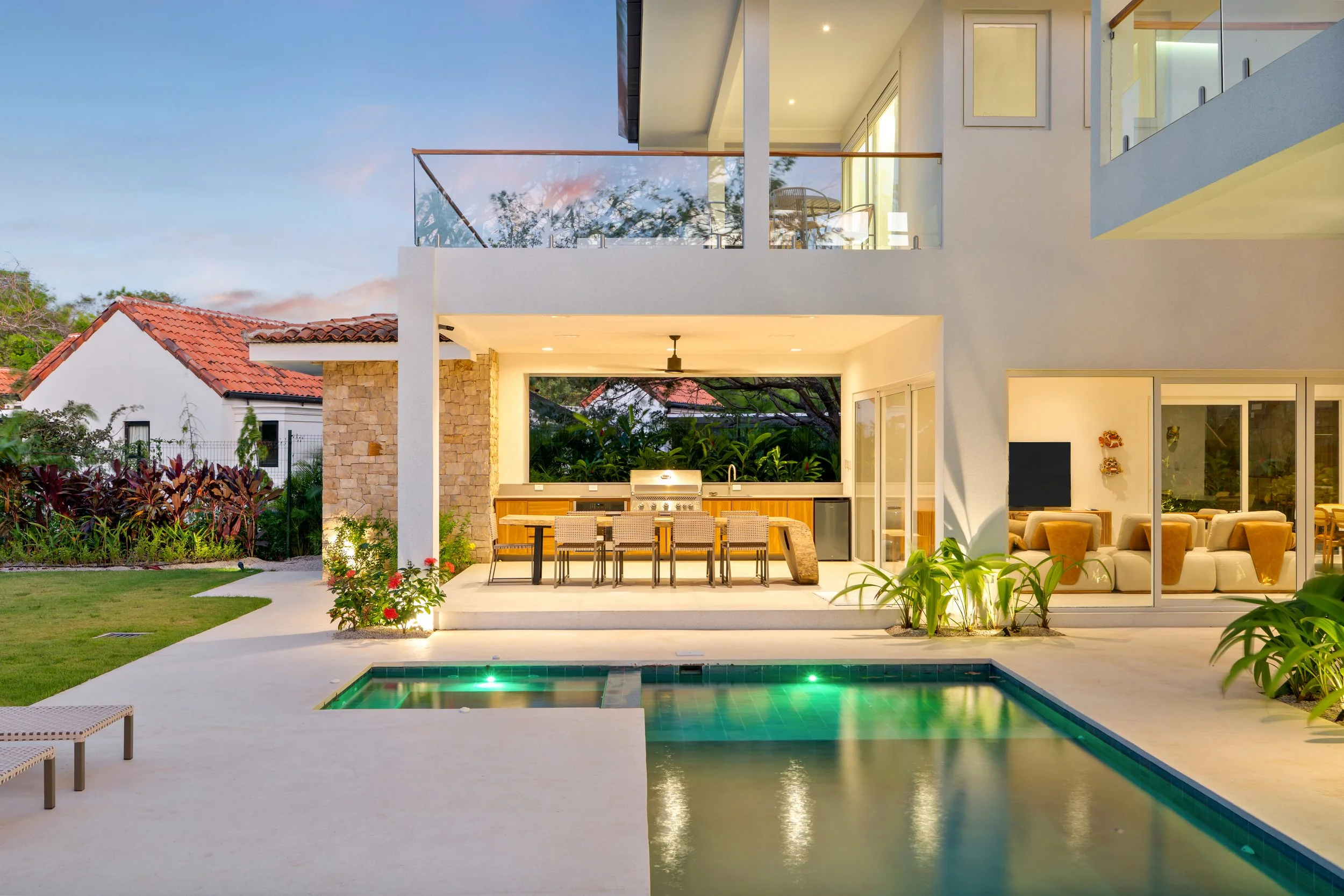Modern two-story house with large glass windows and doors, outdoor dining area, swimming pool, and lush greenery.