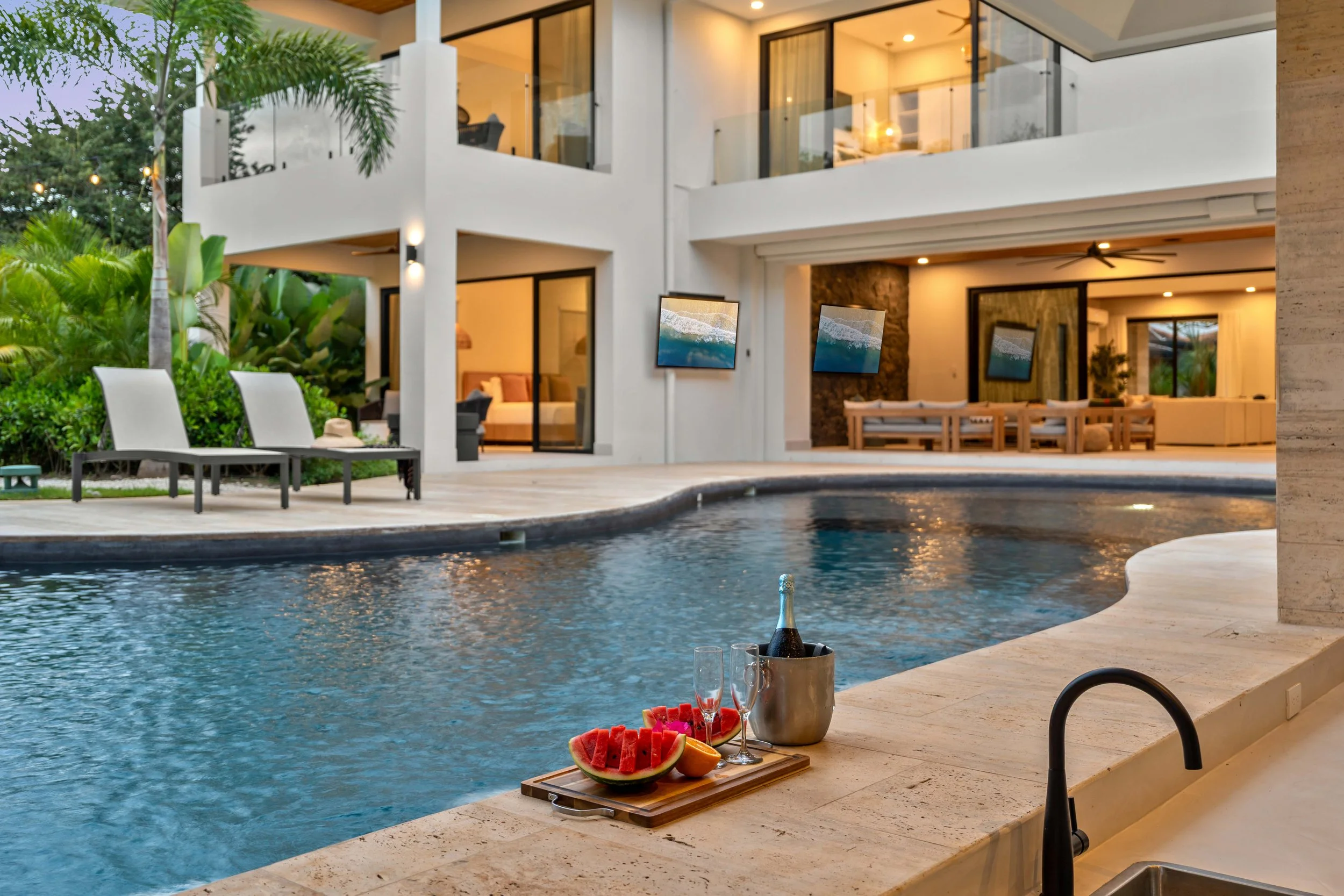 A modern house with large glass windows and a swimming pool in the backyard, with lounge chairs, tropical plants, and a tray with watermelon, orange slices, a bottle of champagne, and champagne flutes.