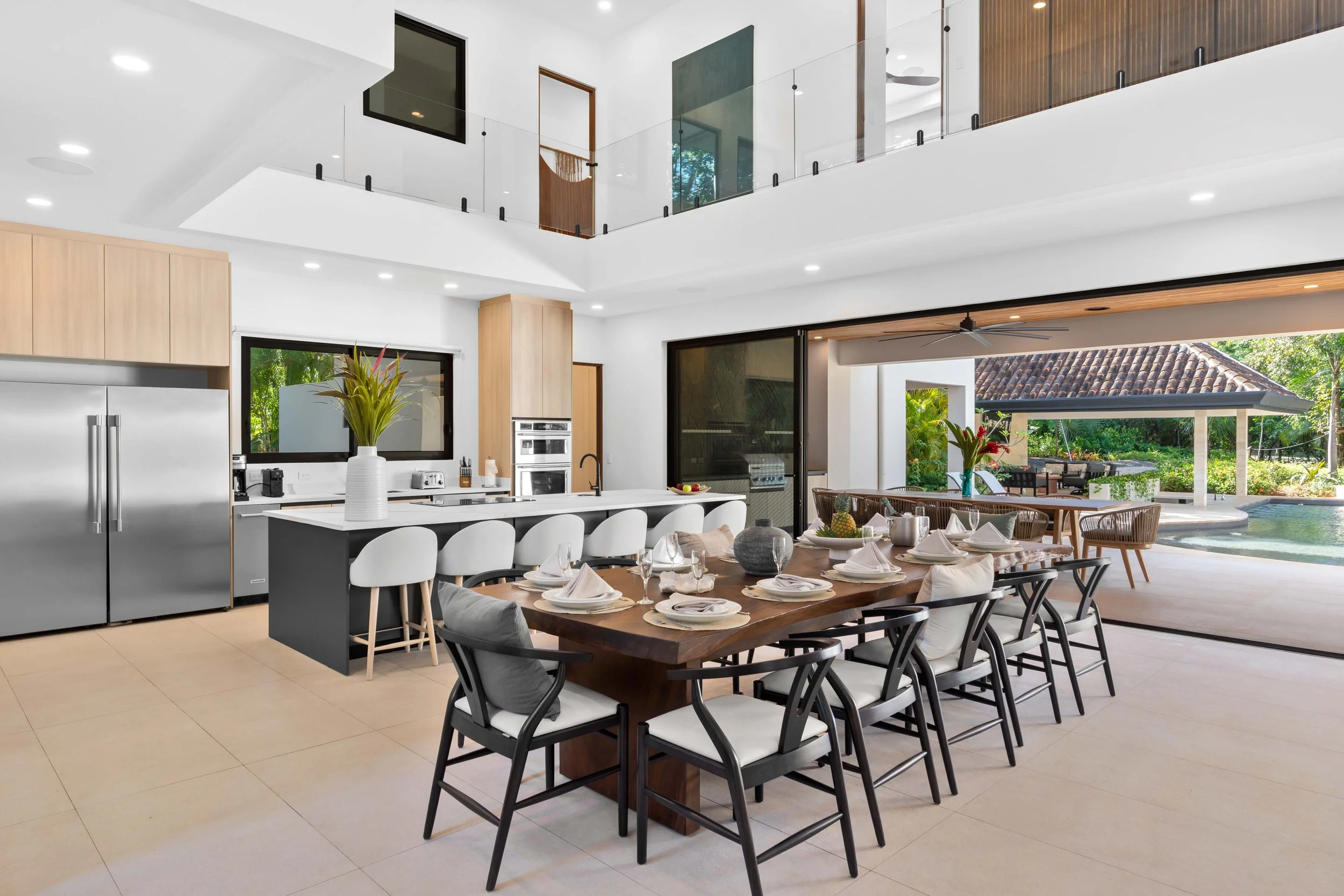 Modern open-concept kitchen and dining area with a large wooden table set for a meal, white chairs, a kitchen island with bar stools, and sliding glass doors leading to an outdoor pool and garden.