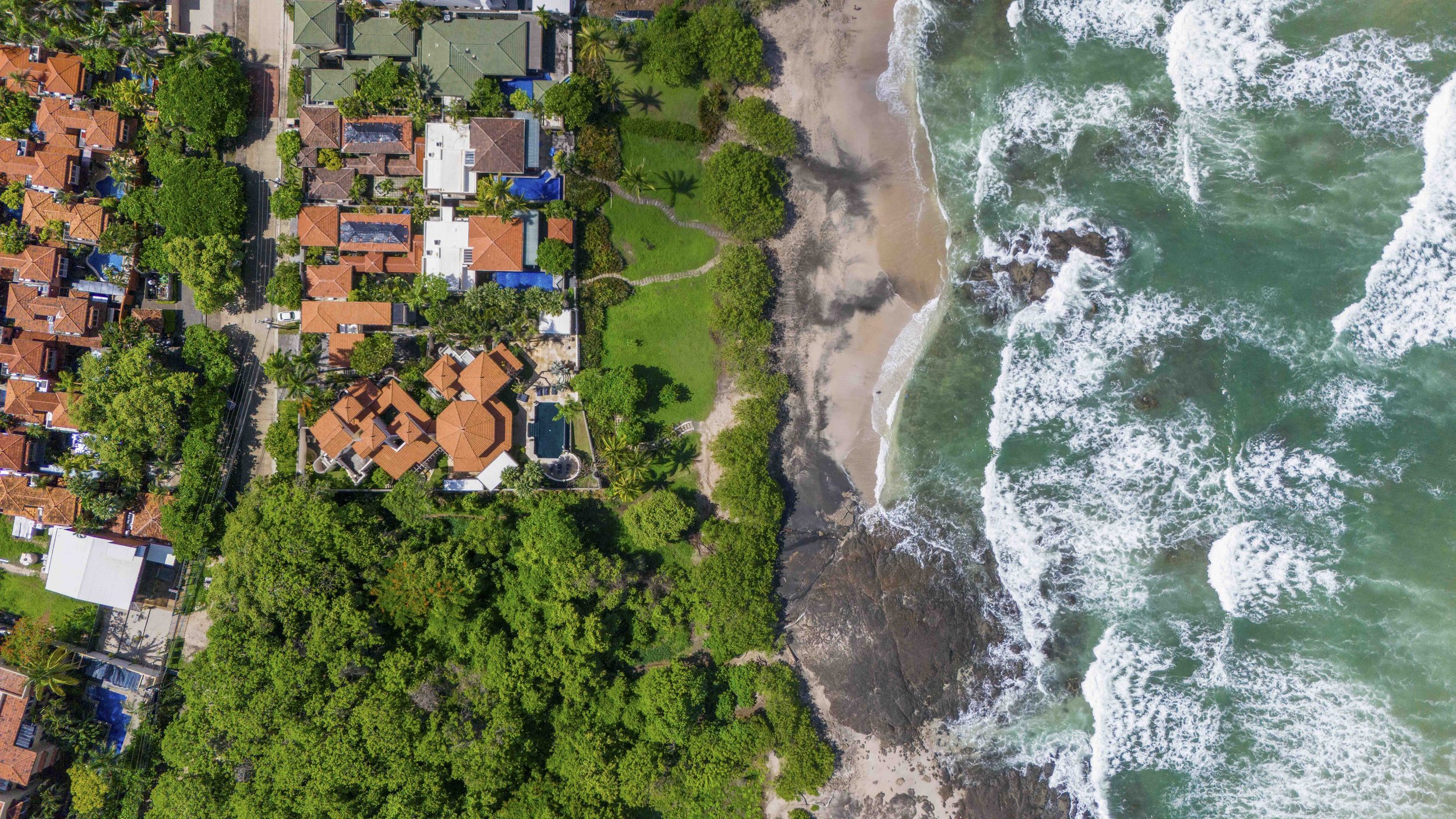 An aerial view in Tamarindo, Costa Rica of a coastal area showing residential homes with red-tiled roofs on the left and lush green trees. The right side features a sandy beach with rocks and ocean waves coming ashore.