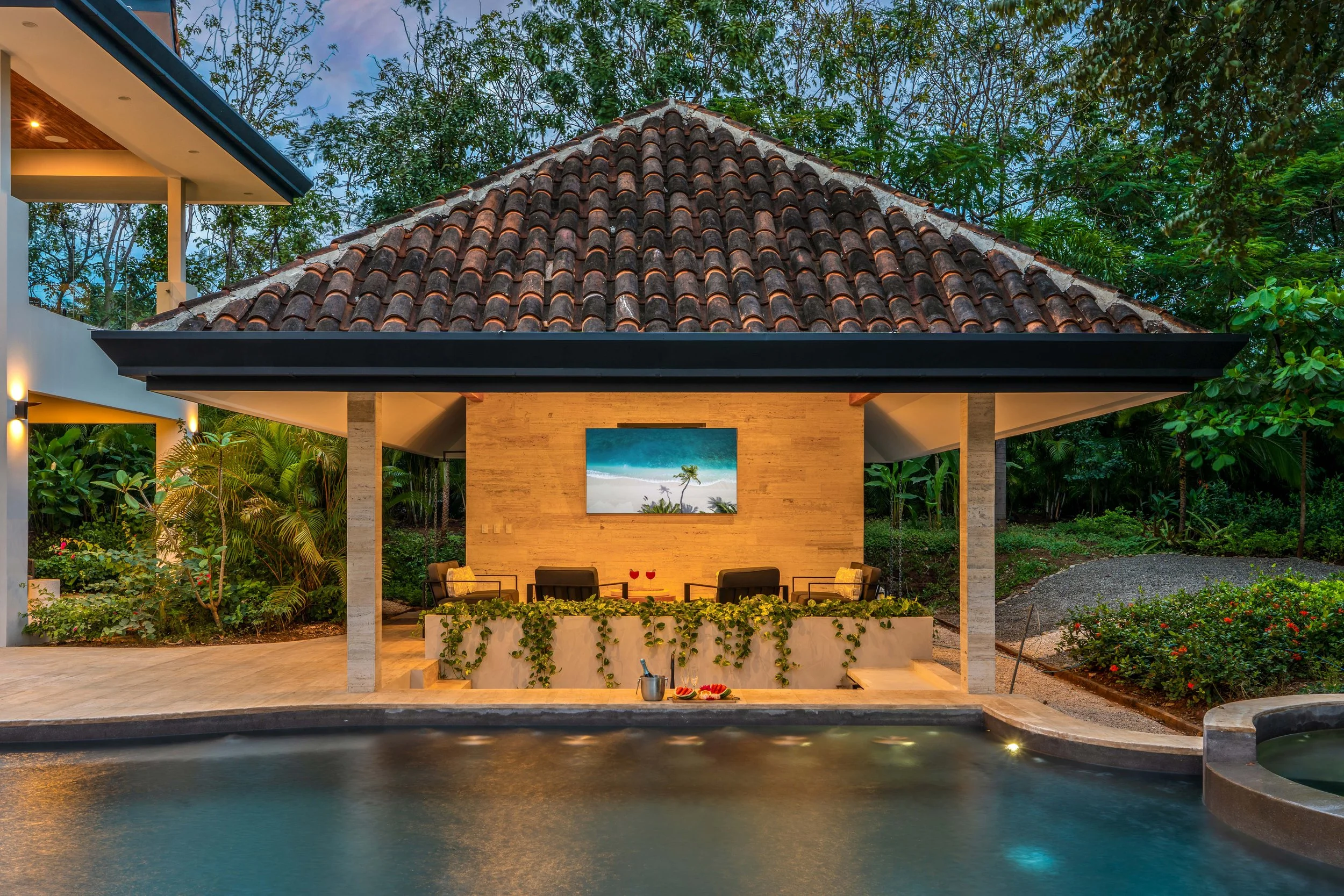 Luxury outdoor pool area with a covered lounge, tropical plants, and a view of a hillside garden at dusk.
